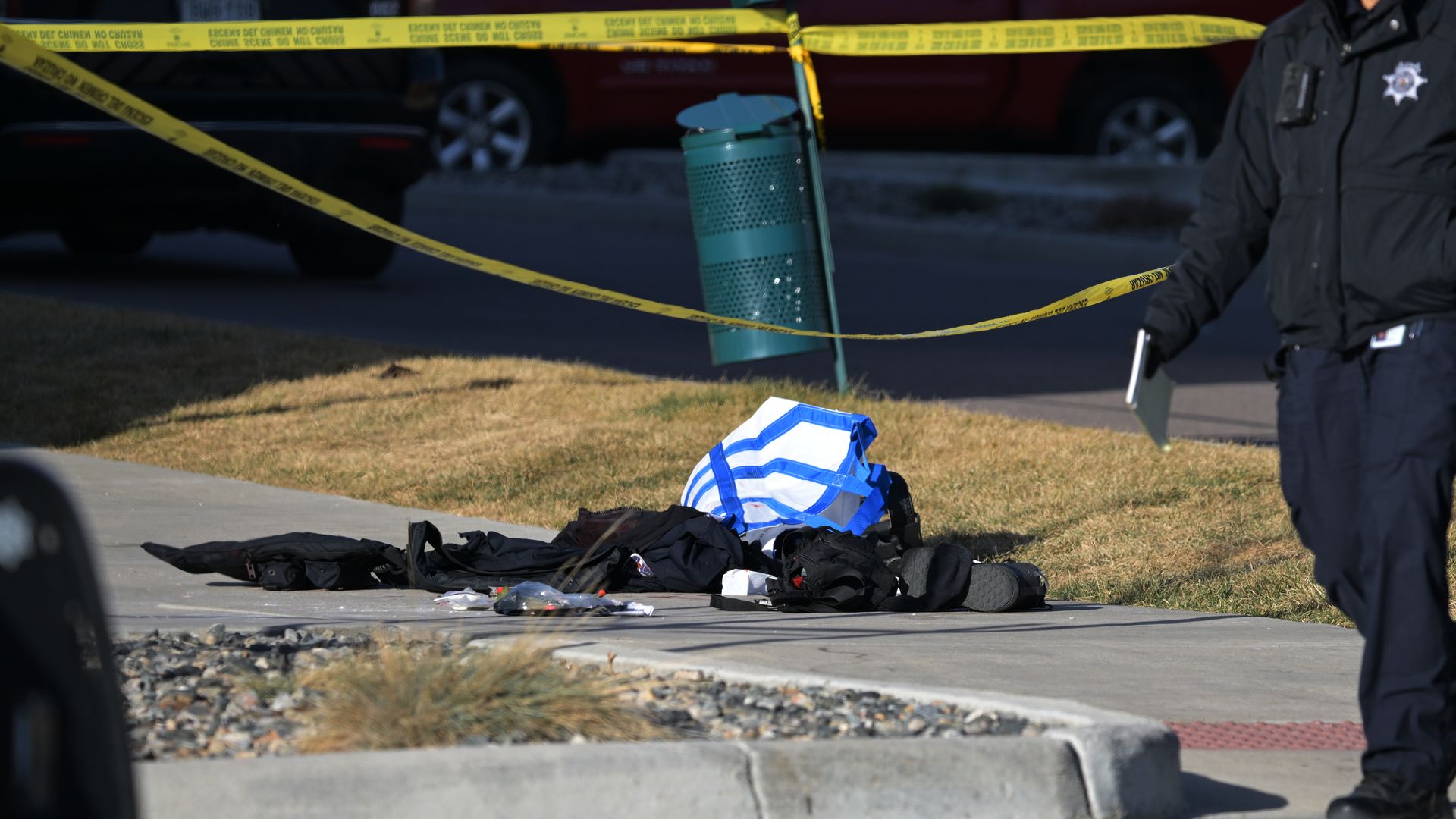 A female officer in a dark jacket stands near a crime scene taped off with yellow caution tape. Debris and clothing lie on the curb, a teal trash can nearby, and police vehicles in the background.