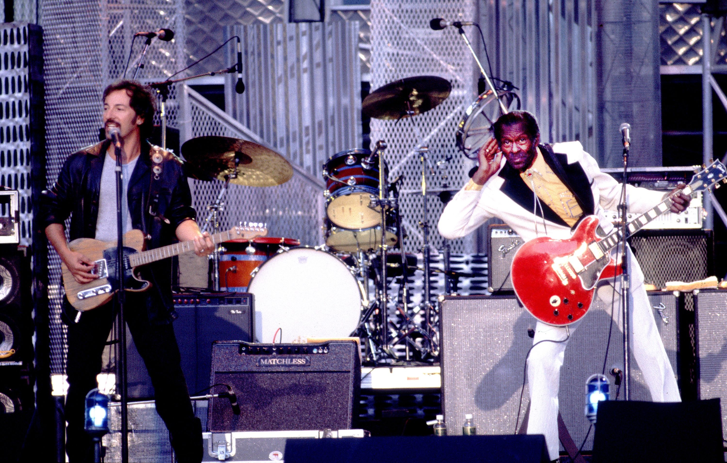 Two male musicians perform on stage: one in a black leather jacket plays a light brown electric guitar and sings, the other in a white suit with a red guitar cups his ear. Drum set in background.
