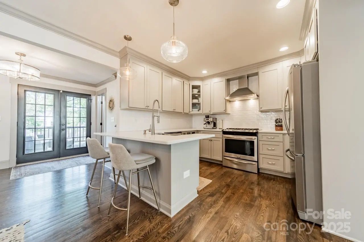 Bright modern kitchen with white cabinets, stainless steel appliances, wooden floor, island with gray stools, glass pendant lights, and black French doors leading outside.