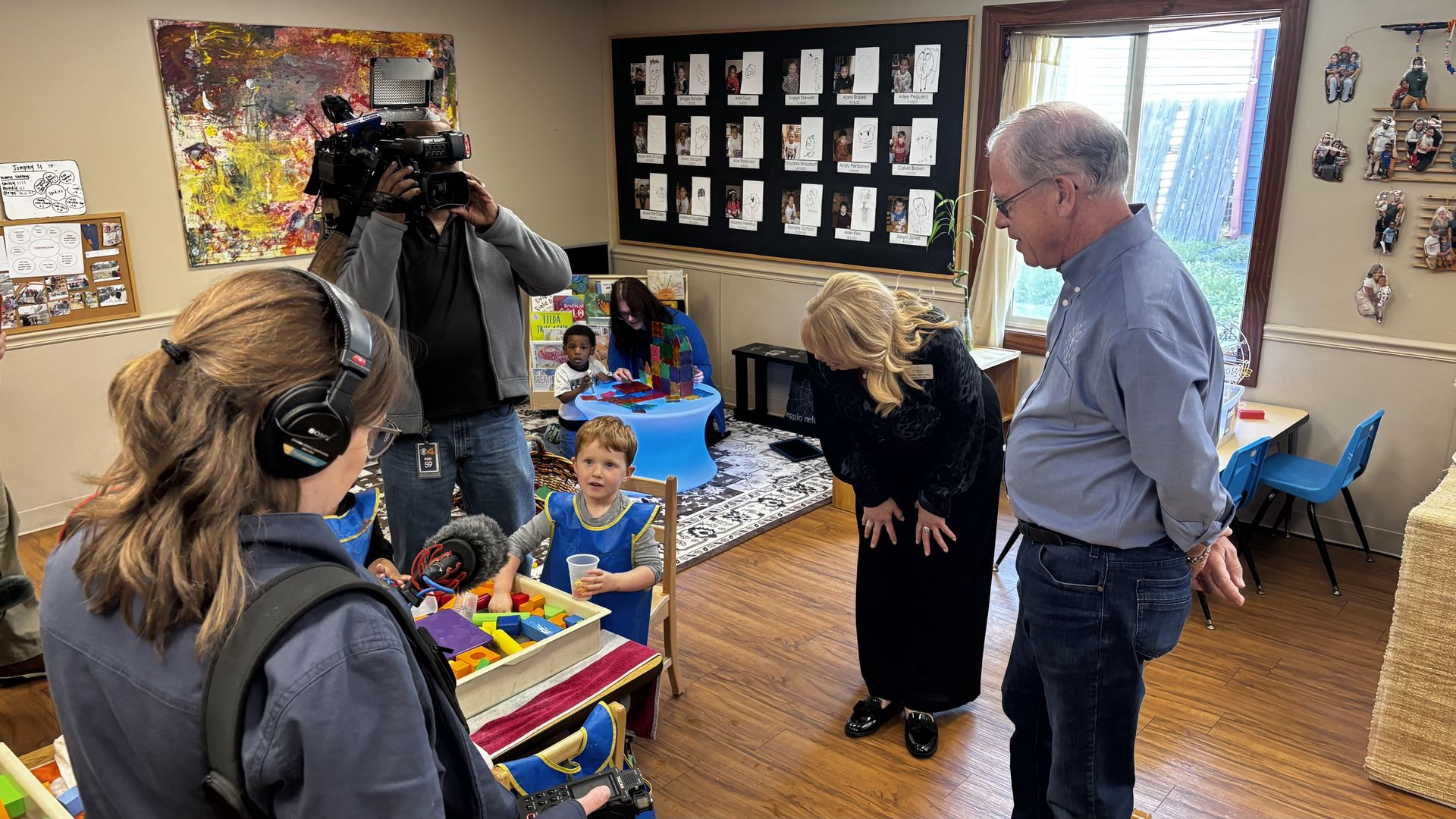 Cameraman films a busy classroom: kids at a blue round table build with colorful blocks, a blonde woman bends over the table, and a man in a blue shirt watches; wall shows student photos.
