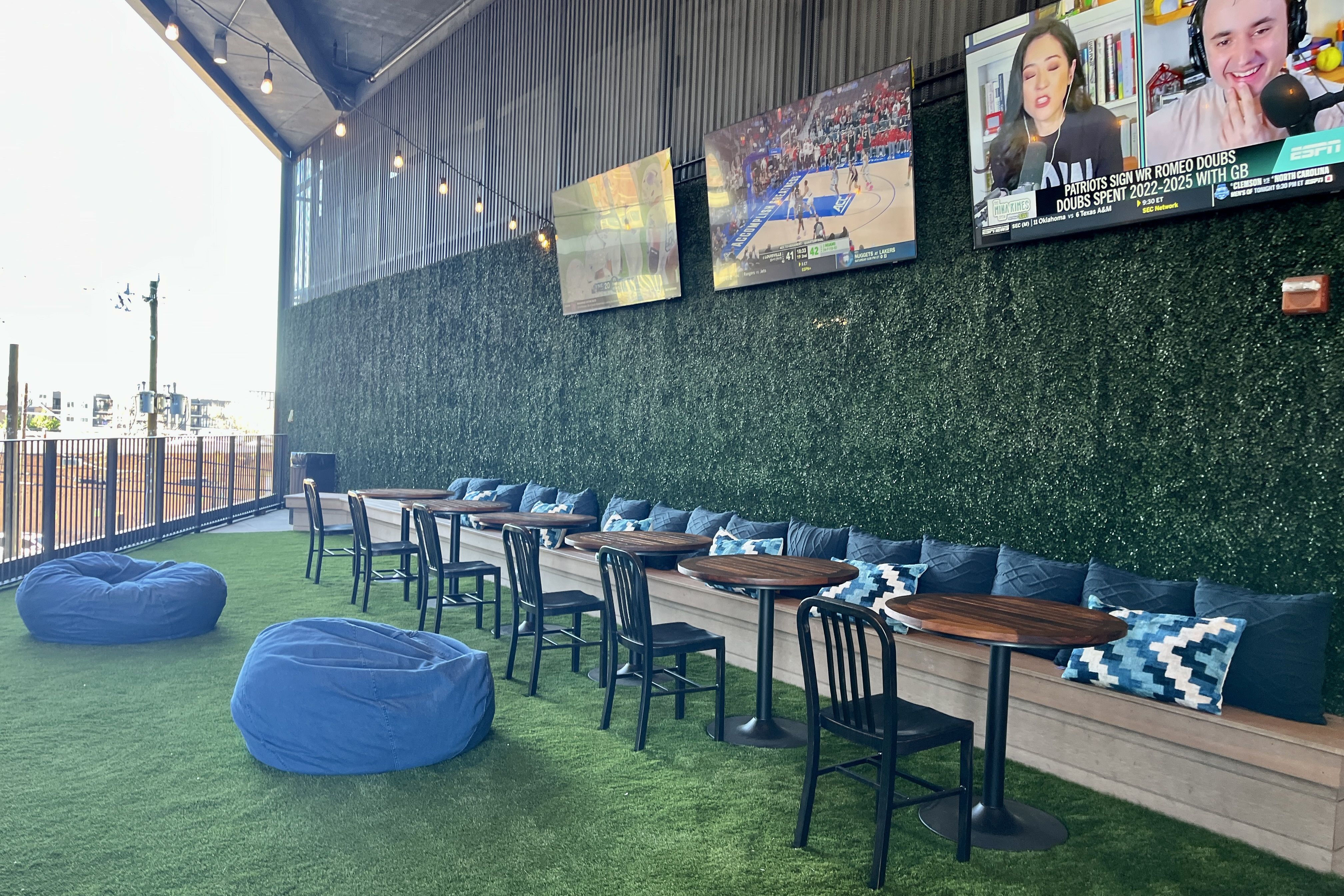Outdoor seating on artificial turf with blue bean bags, round wooden tables, and black chairs along a bench with blue pillows. Hedge wall, multiple TVs showing sports, and string lights overhead.