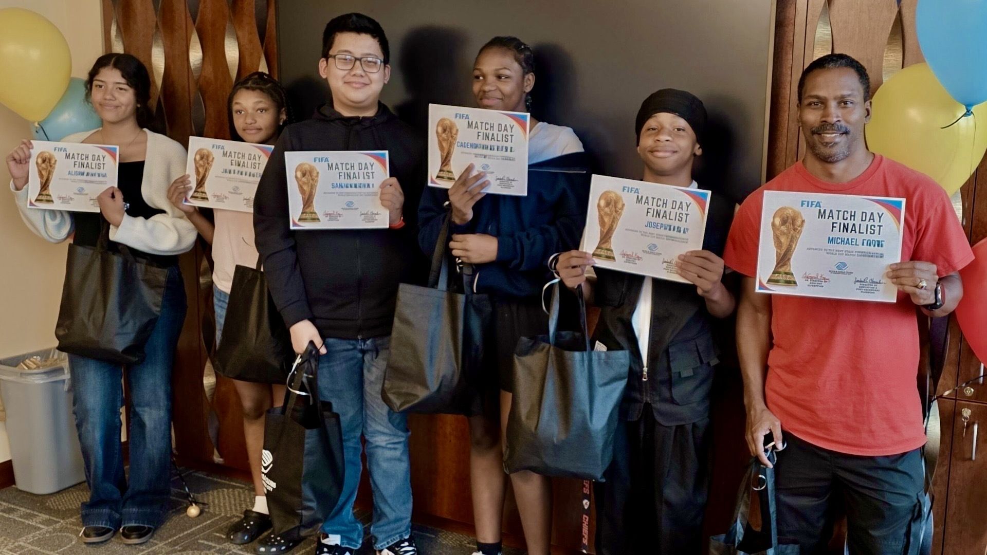 Group of diverse youths and an adult posing indoors, each holding a "FIFA Match Day Finalist" certificate with a trophy image; yellow and blue balloons in the background.