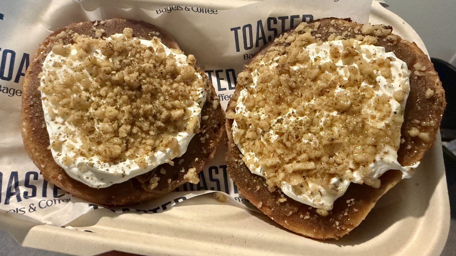 Two toasted bagels with cream cheese and crushed nuts on top, served in a beige cardboard tray lined with paper that reads "TOASTED Bagels & Coffee."