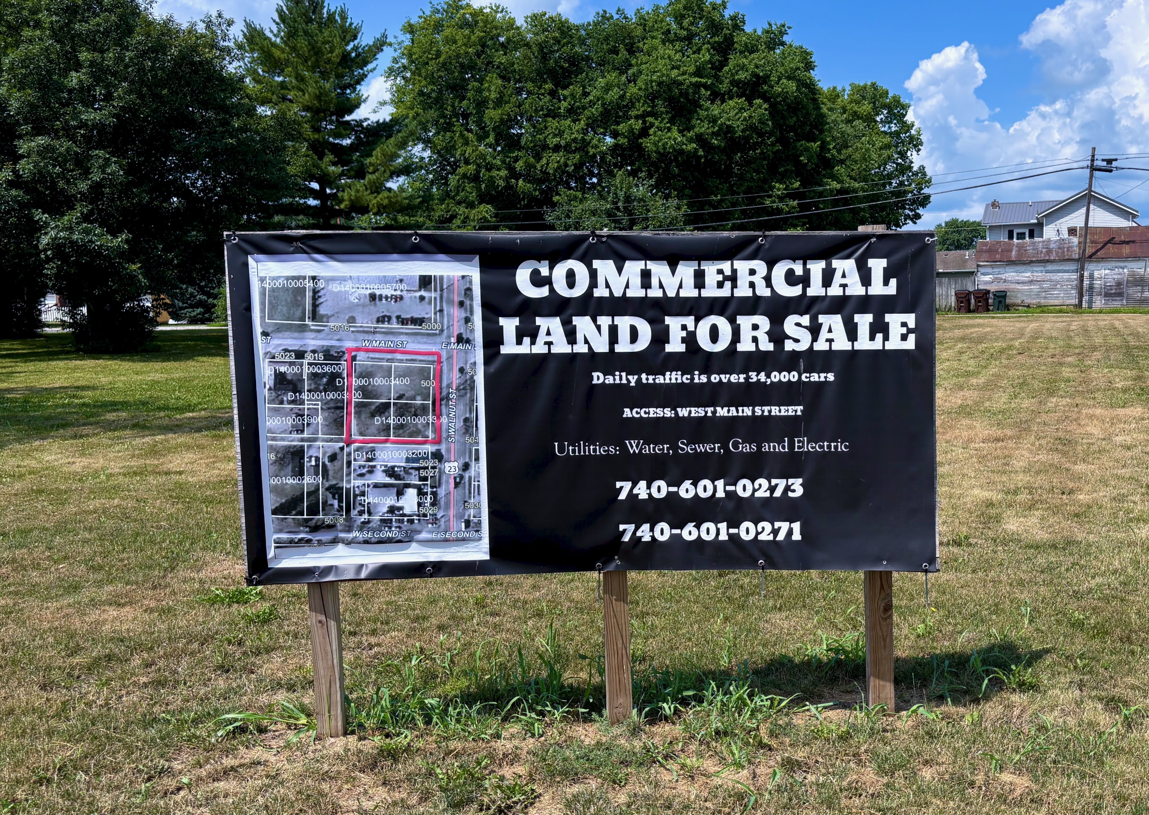 A large black banner reads "COMMERCIAL LAND FOR SALE" with a left-side map, mounted on wooden posts in a grassy lot, with green trees, power lines, and a blue sky with white clouds behind.