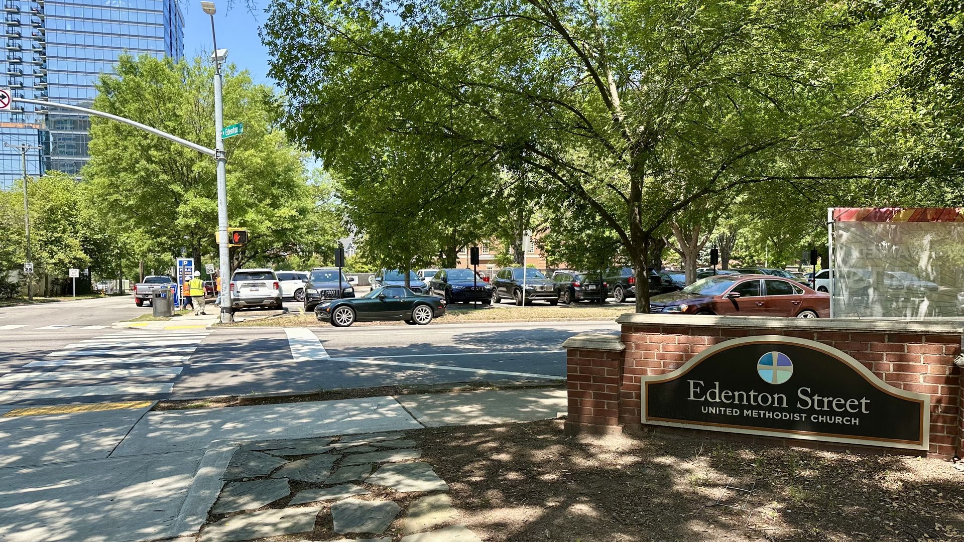 Sunny city street scene with a glass office tower on the left, lush green trees, parked cars, and a brick sign in the foreground reading Edenton Street United Methodist Church.