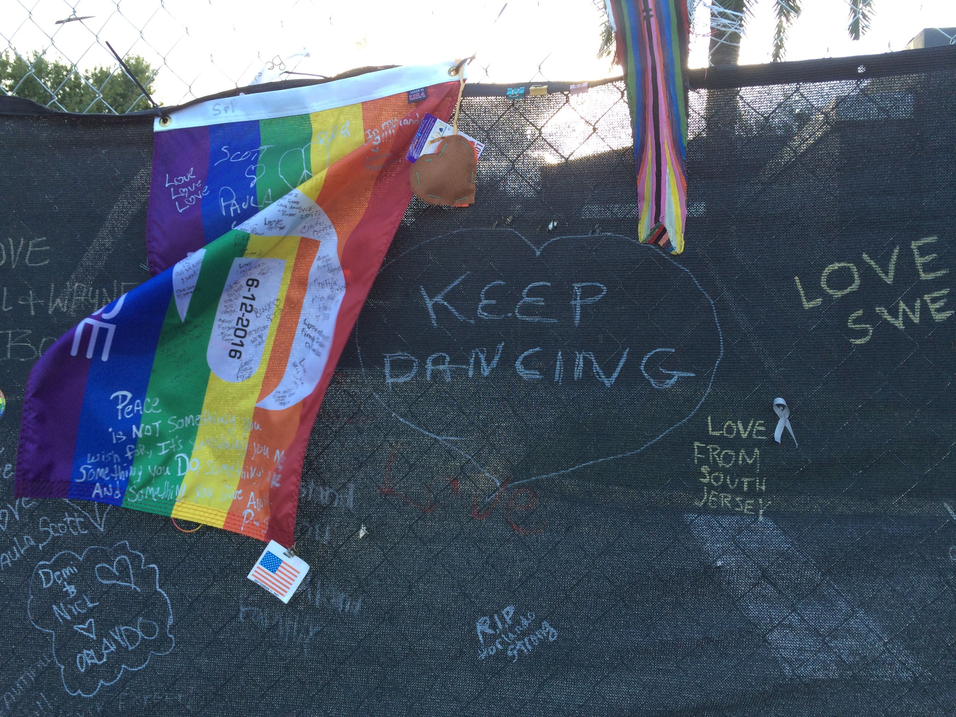 Rainbow Pride flag draped over a chain-link fence with handwritten messages; a brown paper heart hangs from a string, while rainbow ribbons twist nearby and chalked words say "Keep dancing."