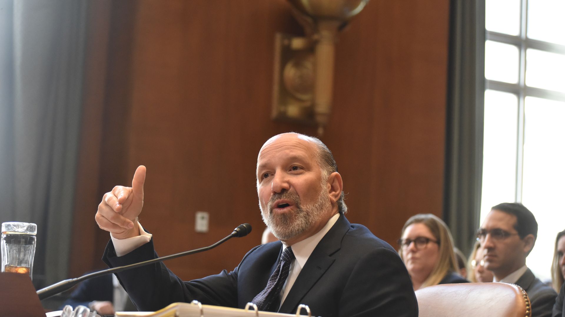 A person in a suit sits in front of a mic and gestures with their hands
