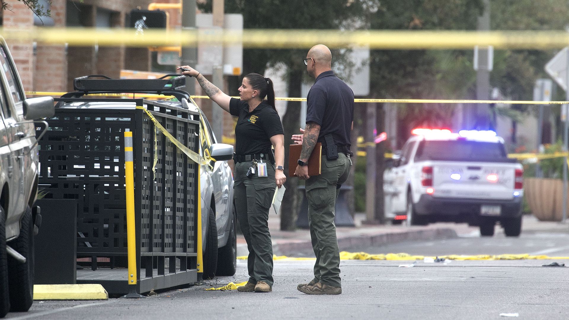 A man and woman, both wearing dark gray pants and dark blue shirts, stand in an area surrounded by yellow crime scene tape as the woman points; a white police vehicle with its lights on can be seen in the background, and cars are parked along a curb. 