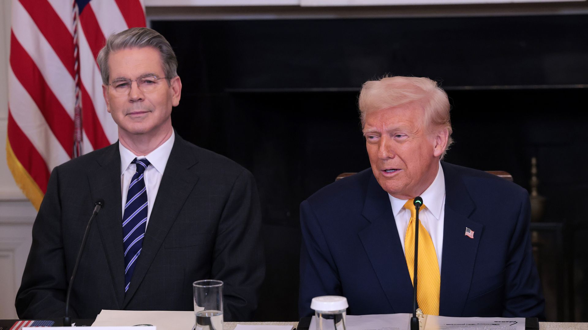 Two men in suits sitting at a table with a US flag behind them. 