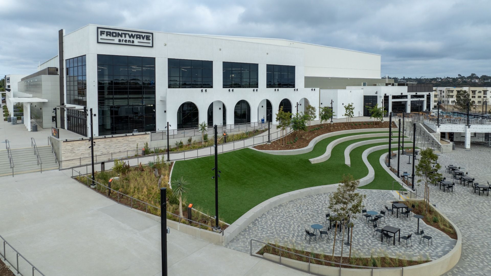 Exterior of Frontwave Arena, a large white building with arched windows, adjacent to a green stepped lawn and an outdoor seating area with tables and chairs under a cloudy sky.