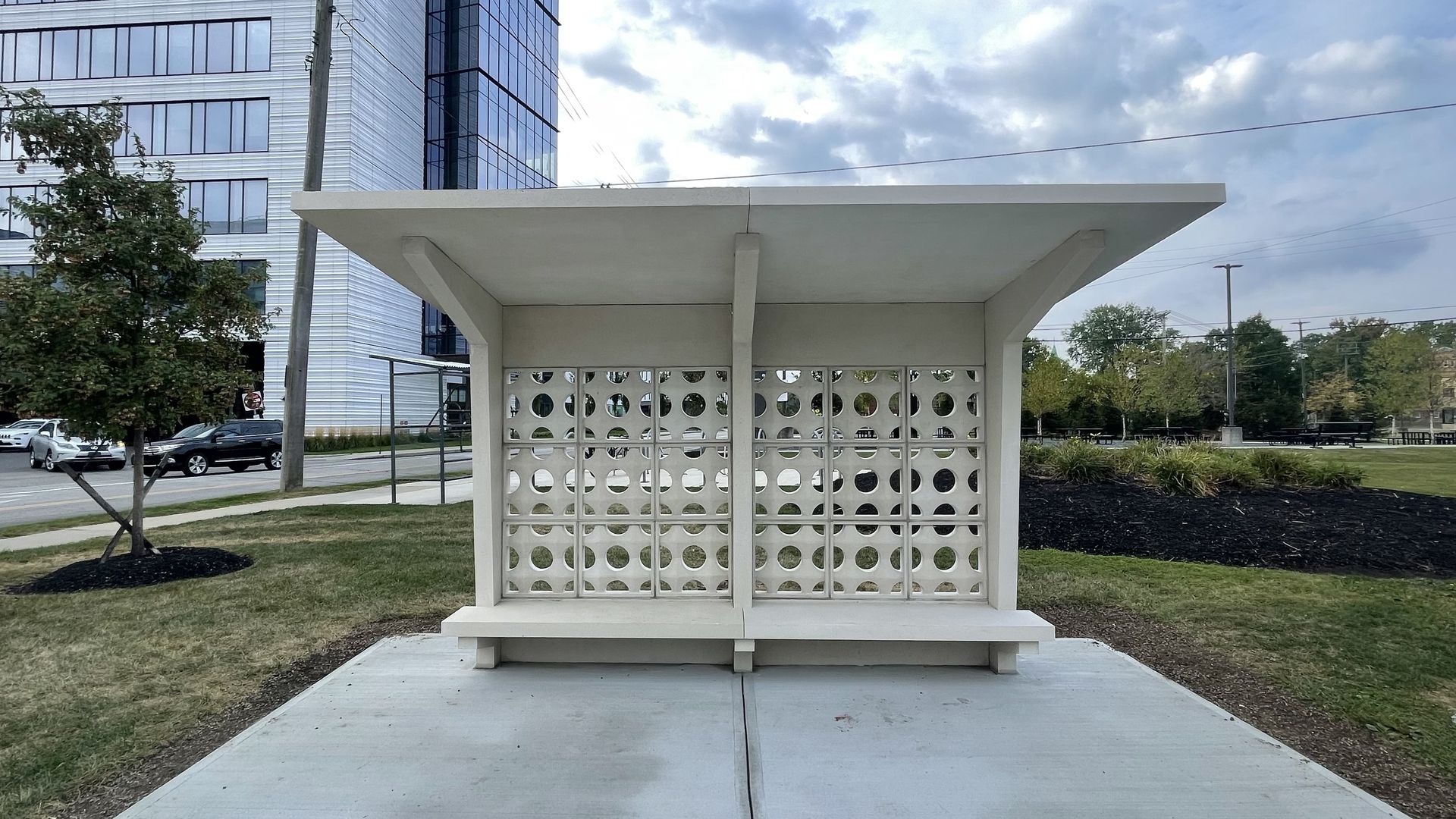 White minimalist public shelter with geometric circular patterns on a beige wall, situated on concrete pavement with grass, trees, a road, and modern glass buildings in the background.