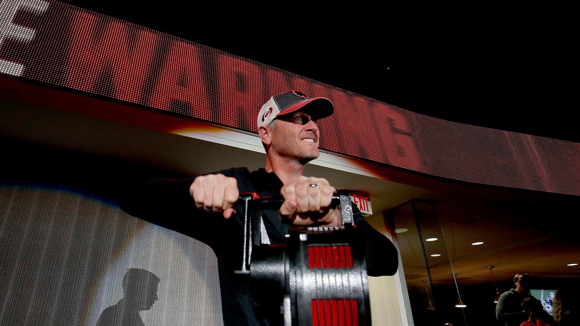A man winds a large black siren, wearing a Carolina Hurricanes hat and grimacing.