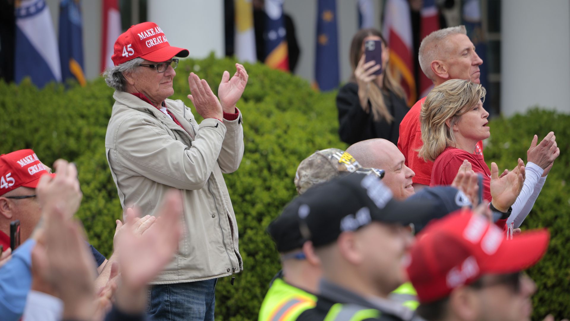 A person in a red hat stands and applauds next to a bush and other applauding people