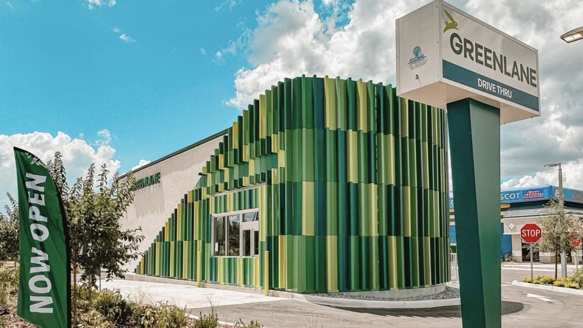 Modern Greenland drive-thru building with green vertical panels, a large sign, and a green "Now Open" flag under a partly cloudy blue sky.