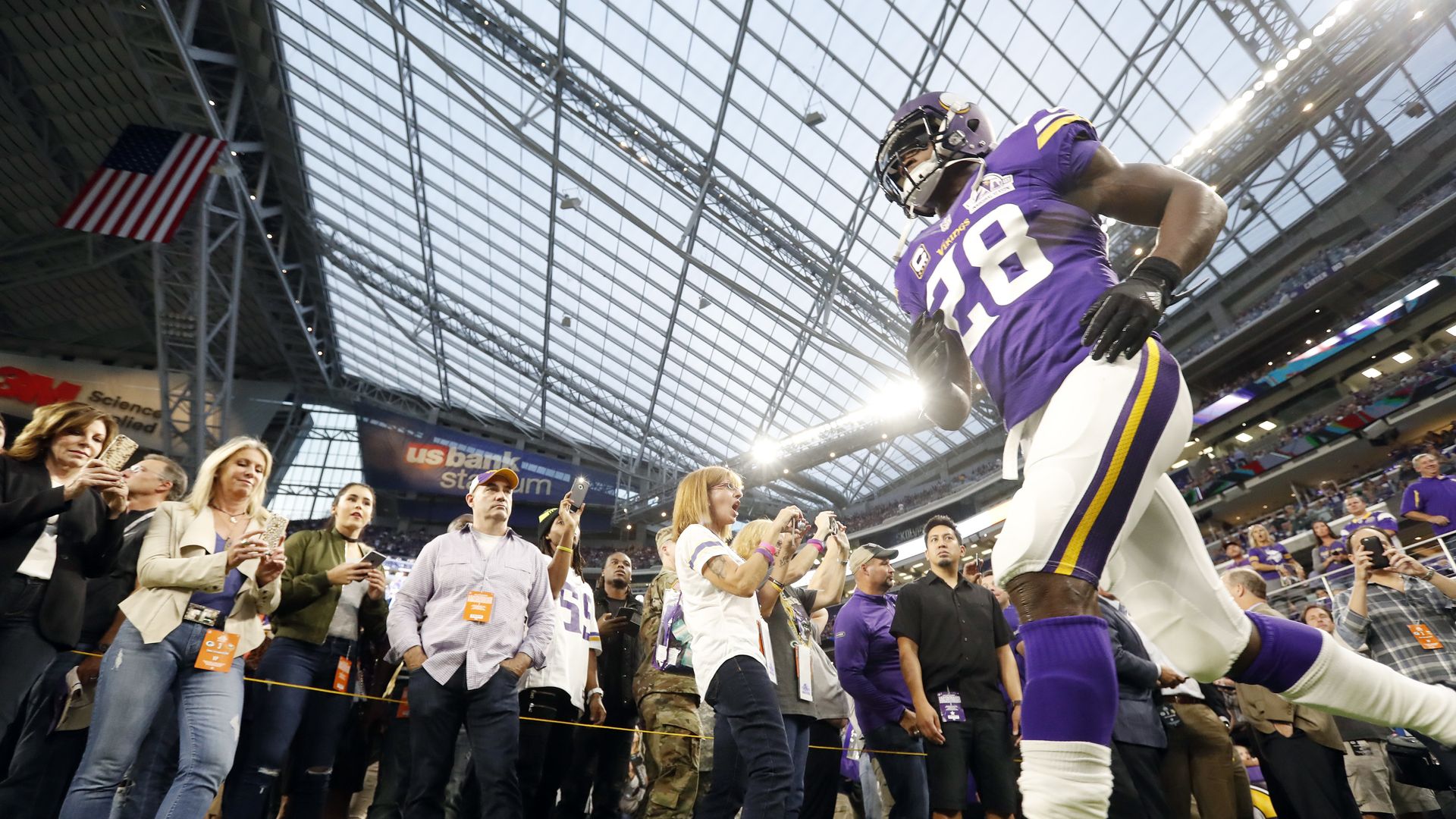 Adrian Peterson walks through a cheering crowd on the field inside a glass-roof stadium, fans taking photos as an American flag hangs overhead.