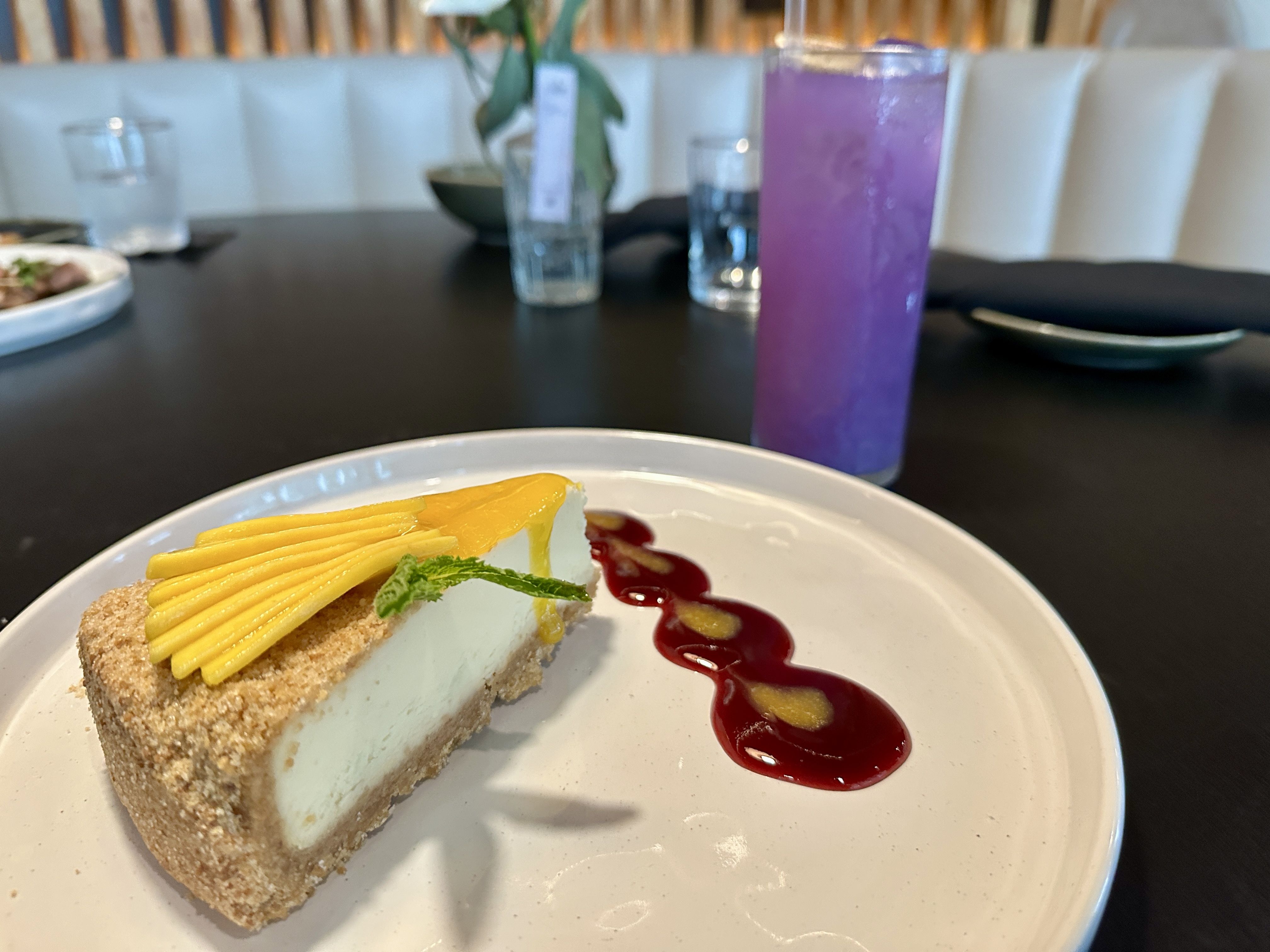 LILA. Slice of pale blue cake with a crumb crust, topped with mango slices and a mint leaf, on a white plate with red berry sauce. A blurred purple drink and glassware sit on a dark table in the background.