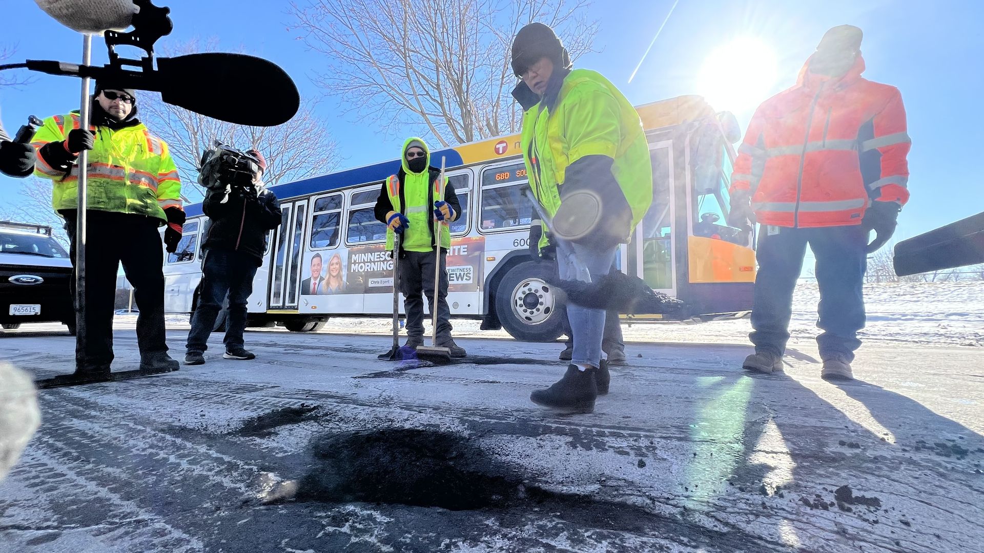 Road workers in bright yellow and orange jackets repairing a pothole on a snowy street near a bus with a winter scene and clear blue sky in the background.