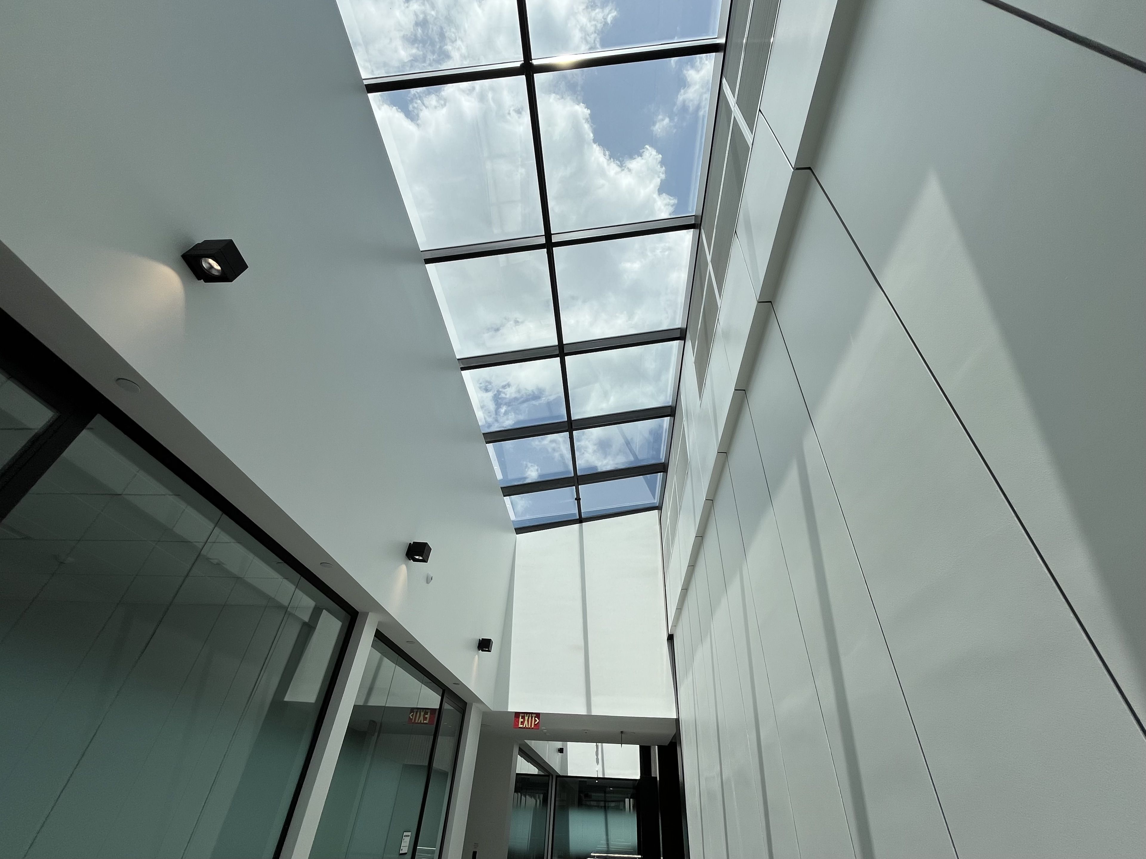 Bright modern atrium with a glass skylight grid overhead revealing blue sky and clouds. White paneled walls, glass doors along a corridor, and red EXIT signs near the far end.