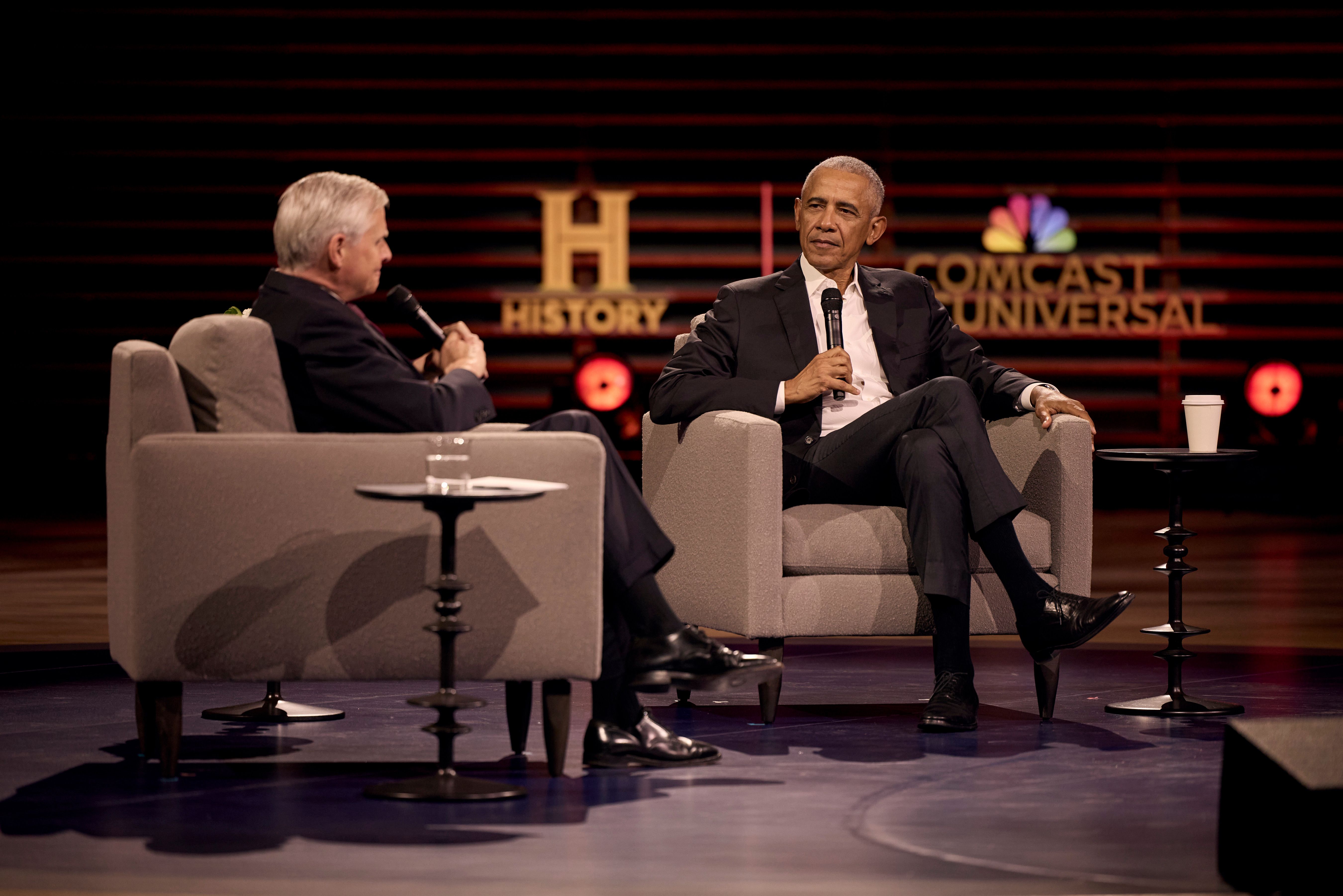 Two men in dark suits sit in grey armchairs on stage, each with a microphone. A water glass sits left; a coffee cup sits right. Red backdrop displays HISTORY and COMCAST UNIVERSAL logos.