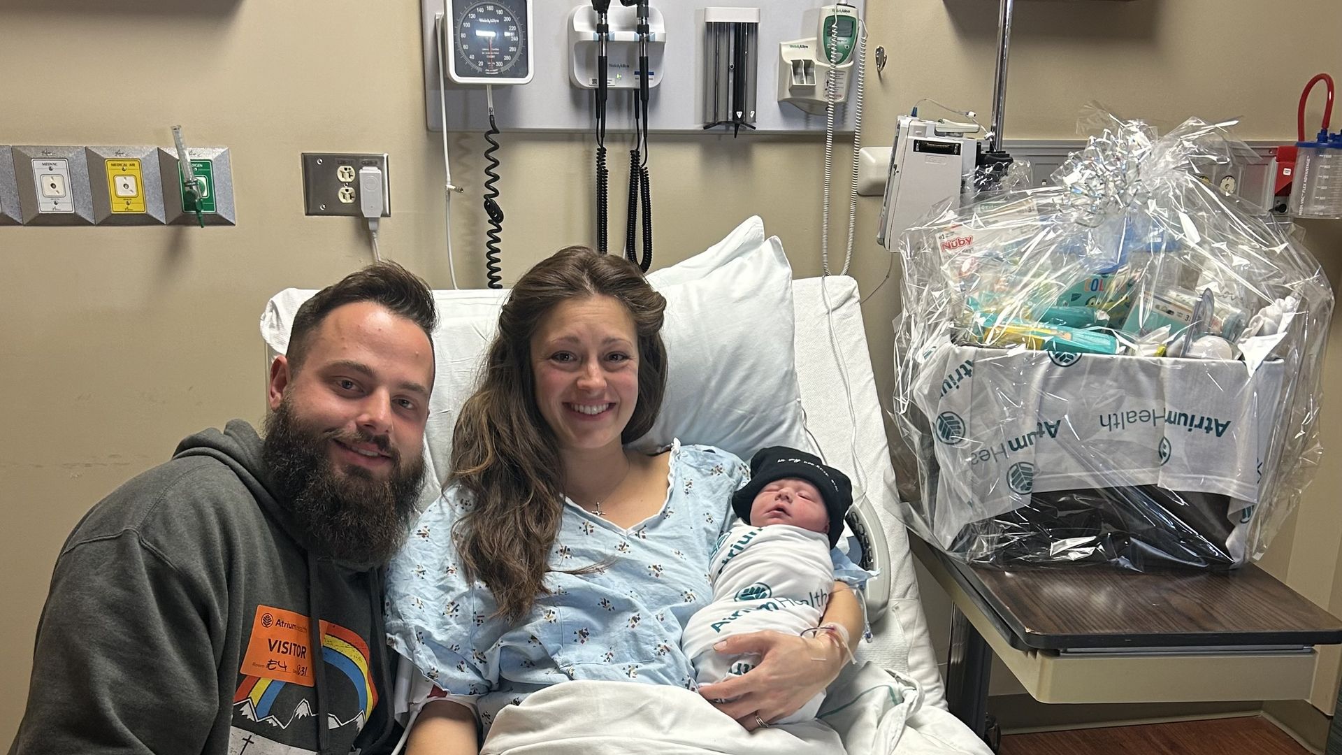 A photo showing a father with a beard, and a mother in a hospital bed holding a newborn baby next to a gift basket. 