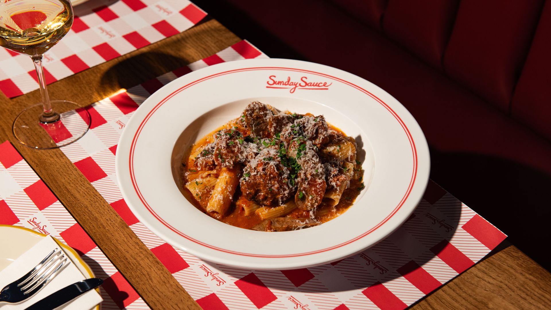 White bowl of rigatoni pasta with red meat sauce, grated cheese, and chopped herbs, on a wooden table with red and white checkered placemats and a glass of white wine nearby.