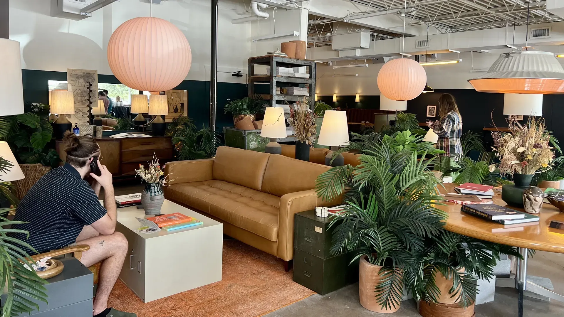 Open-plan lounge with abundant greenery, a tan leather sofa, and pink hanging lamps. A man on the left talks on a phone; a plaid-wearing woman on the right uses a device amid plants.