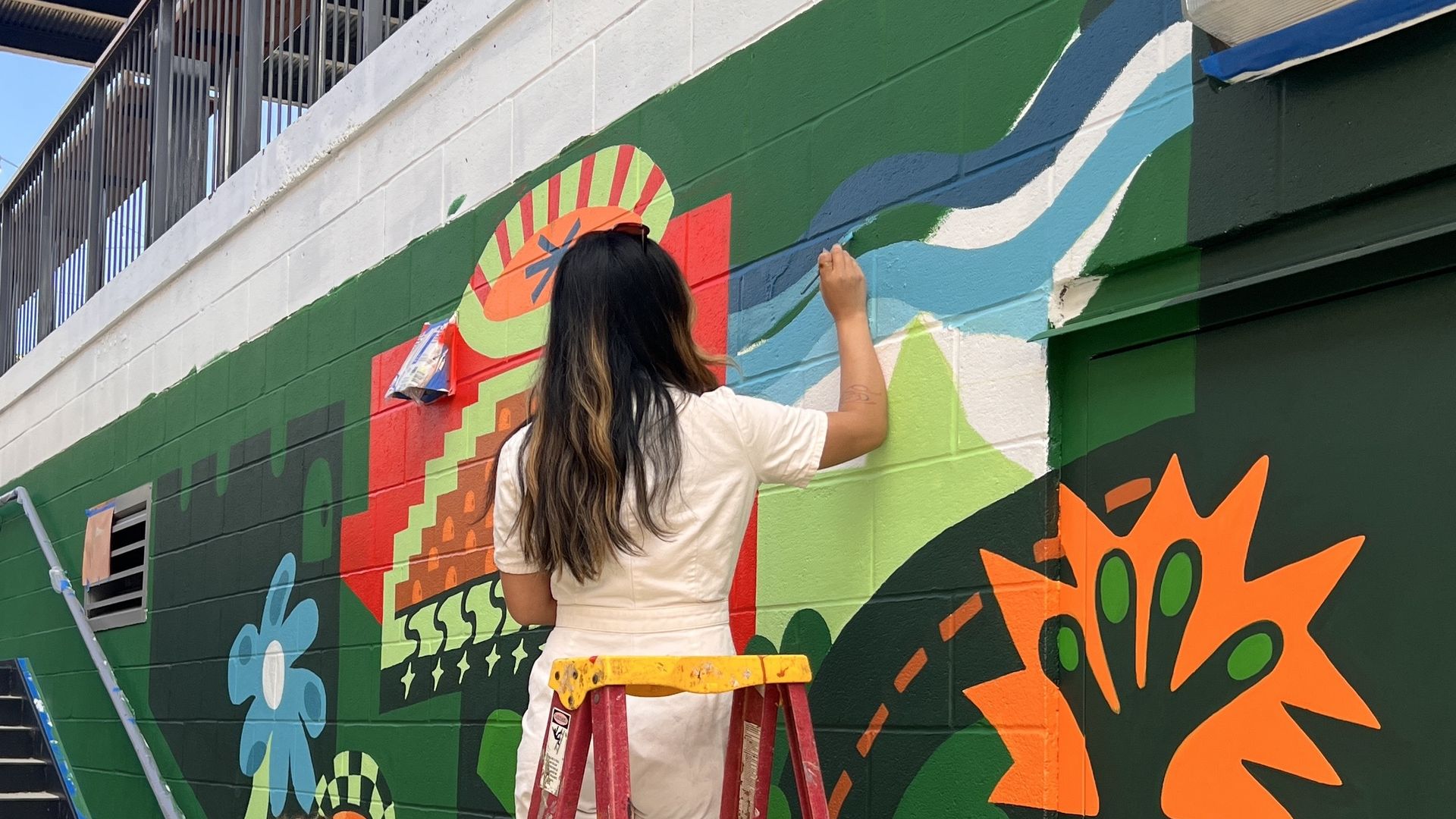 Two artists paint a colorful mural on a green wall beside a stairway. A woman on a red ladder adds orange shapes while another tattooed artist arranges paints on the ground.