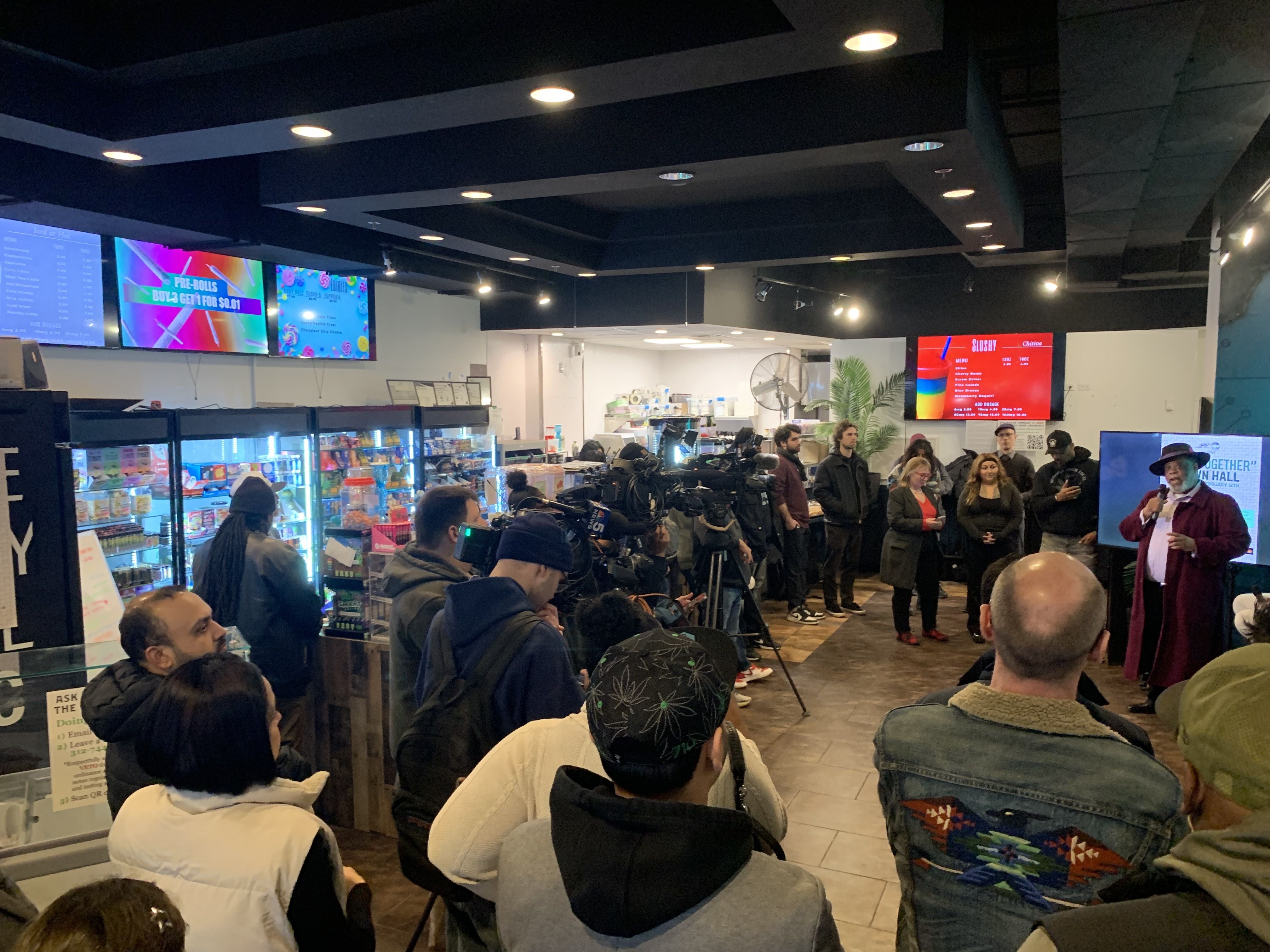 A speaker in a burgundy coat and hat addresses a crowd inside a store with bright fridge displays, cameras, and screens showing menus and event info around the room.