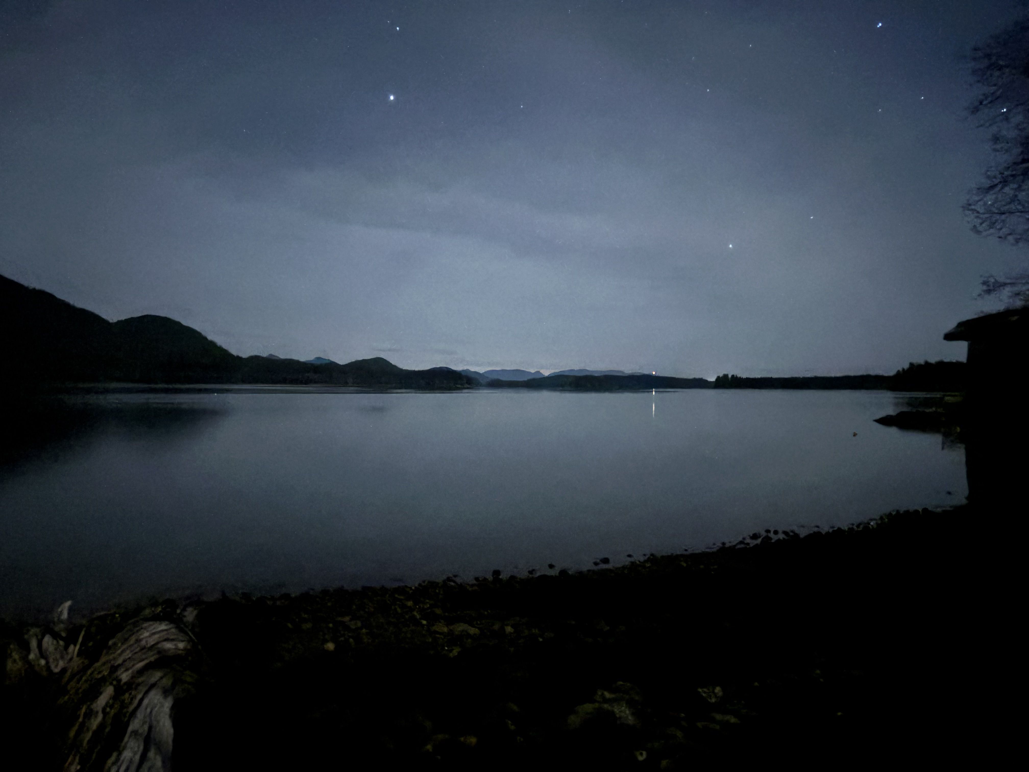 Calm night lake scene with dark silhouettes of distant hills, a mostly clear starry sky, faint clouds, and a small light reflection on the water near the horizon.