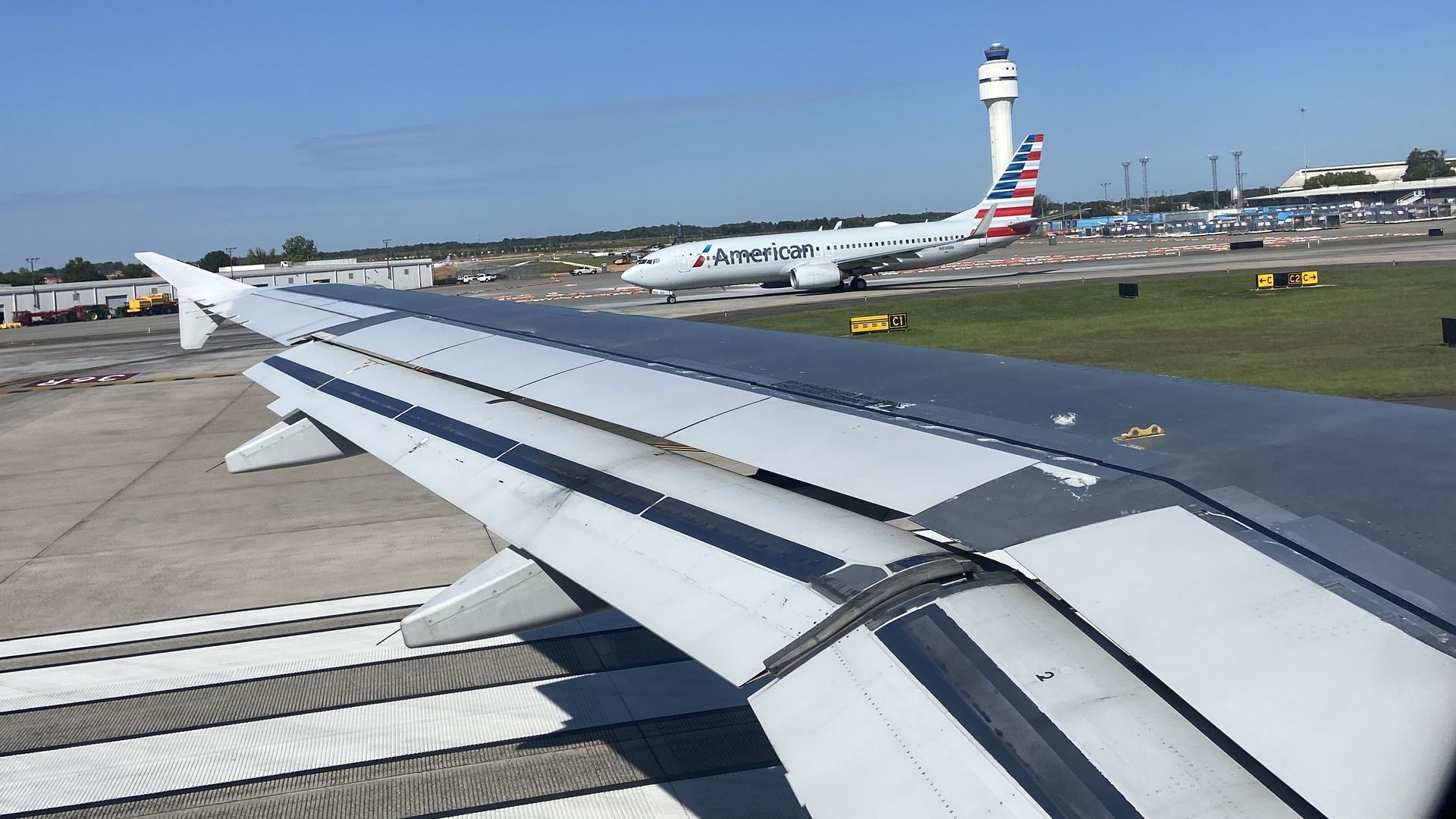 View from airplane window showing wing and runway, with an American Airlines plane and airport control tower in the background under a clear blue sky in Charlotte. 