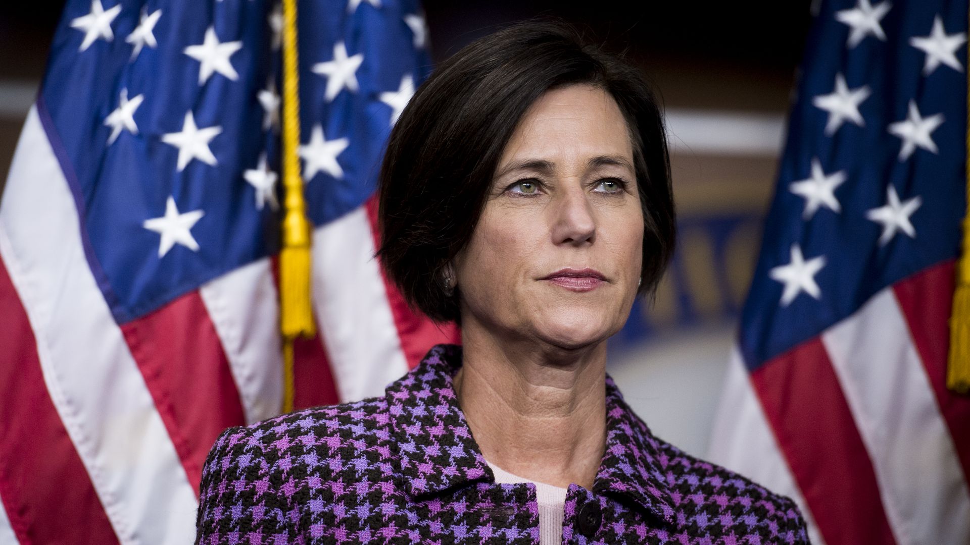 Mimi Walters in a purple plaid jacket standing in front of two American flags