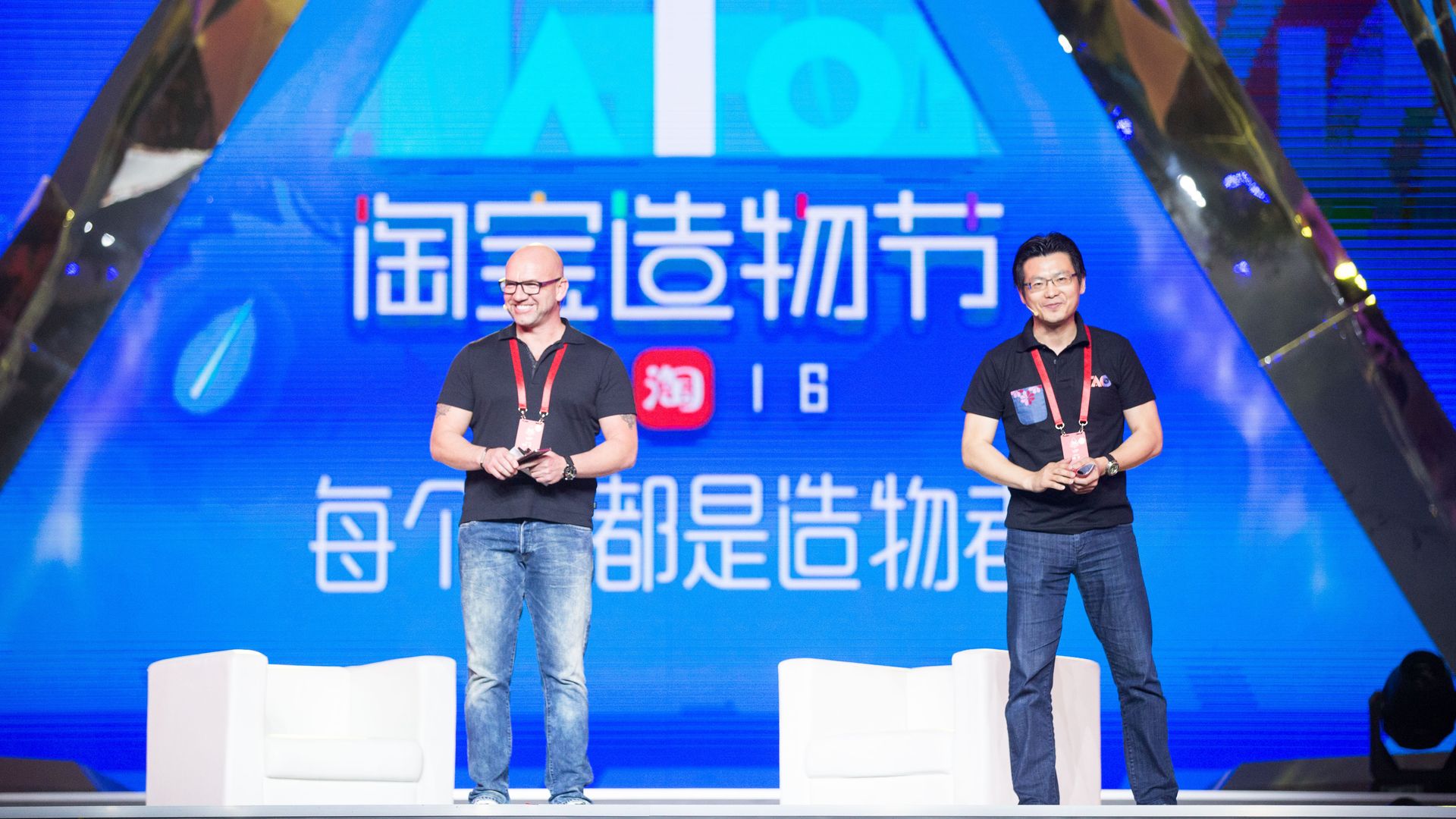 Two men in back T-shirts and jeans stand on a stage with two white chairs. 