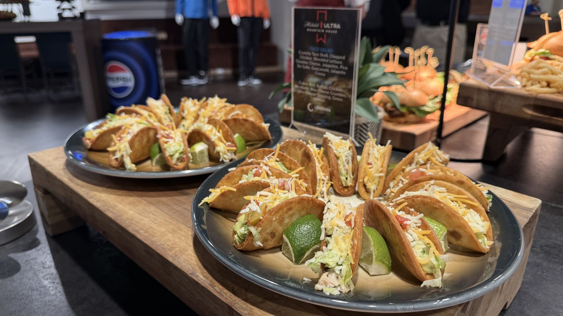 Blurry restaurant scene: plates of soft tacos filled with meat, lettuce, and shredded cheese on a wooden table; lime wedges visible; blurred background shows jackets on mannequins.