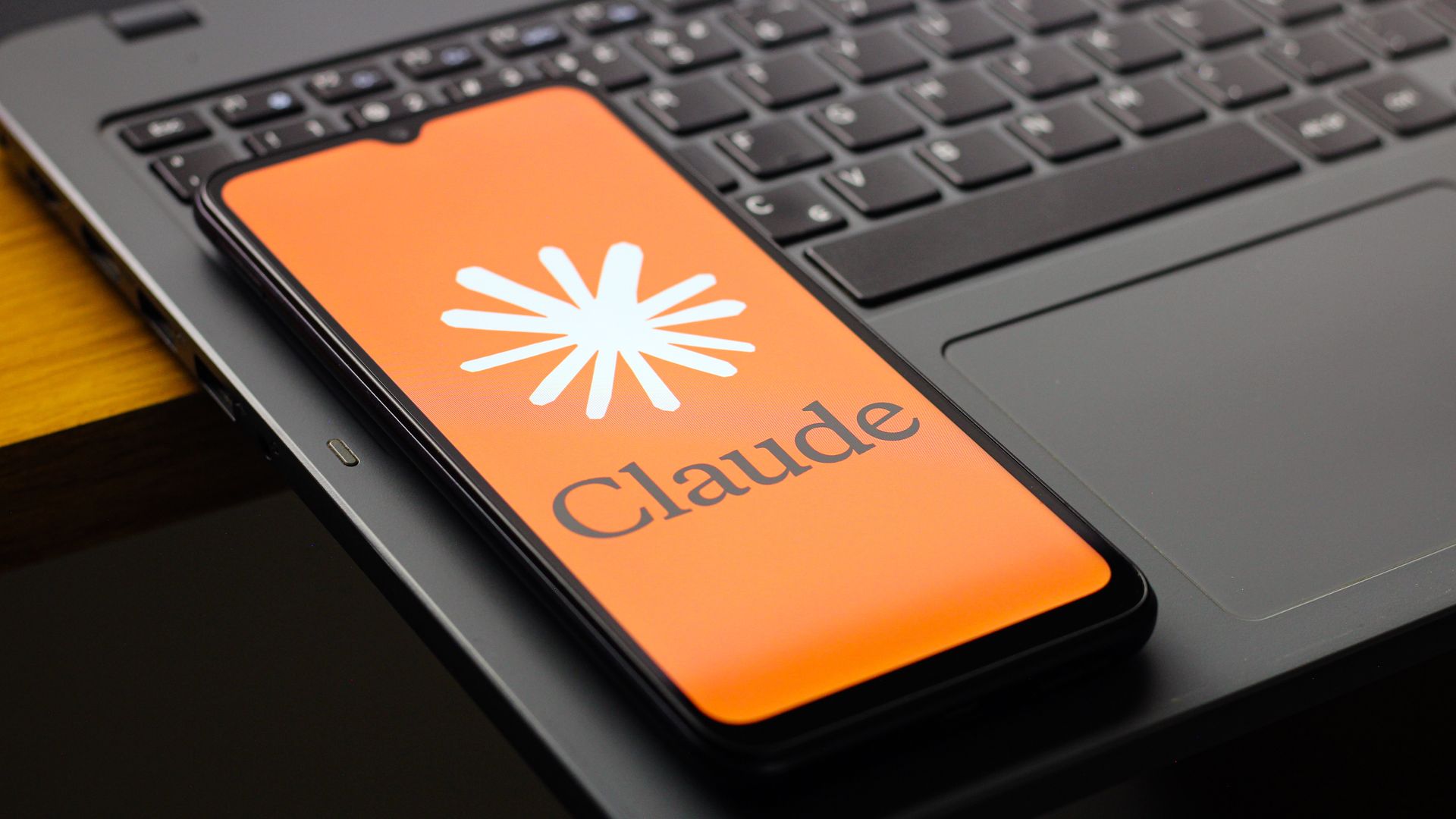 Close-up of a laptop keyboard with a smartphone resting on it. The phone screen is bright orange with a white starburst logo and part of the word Claude visible.
