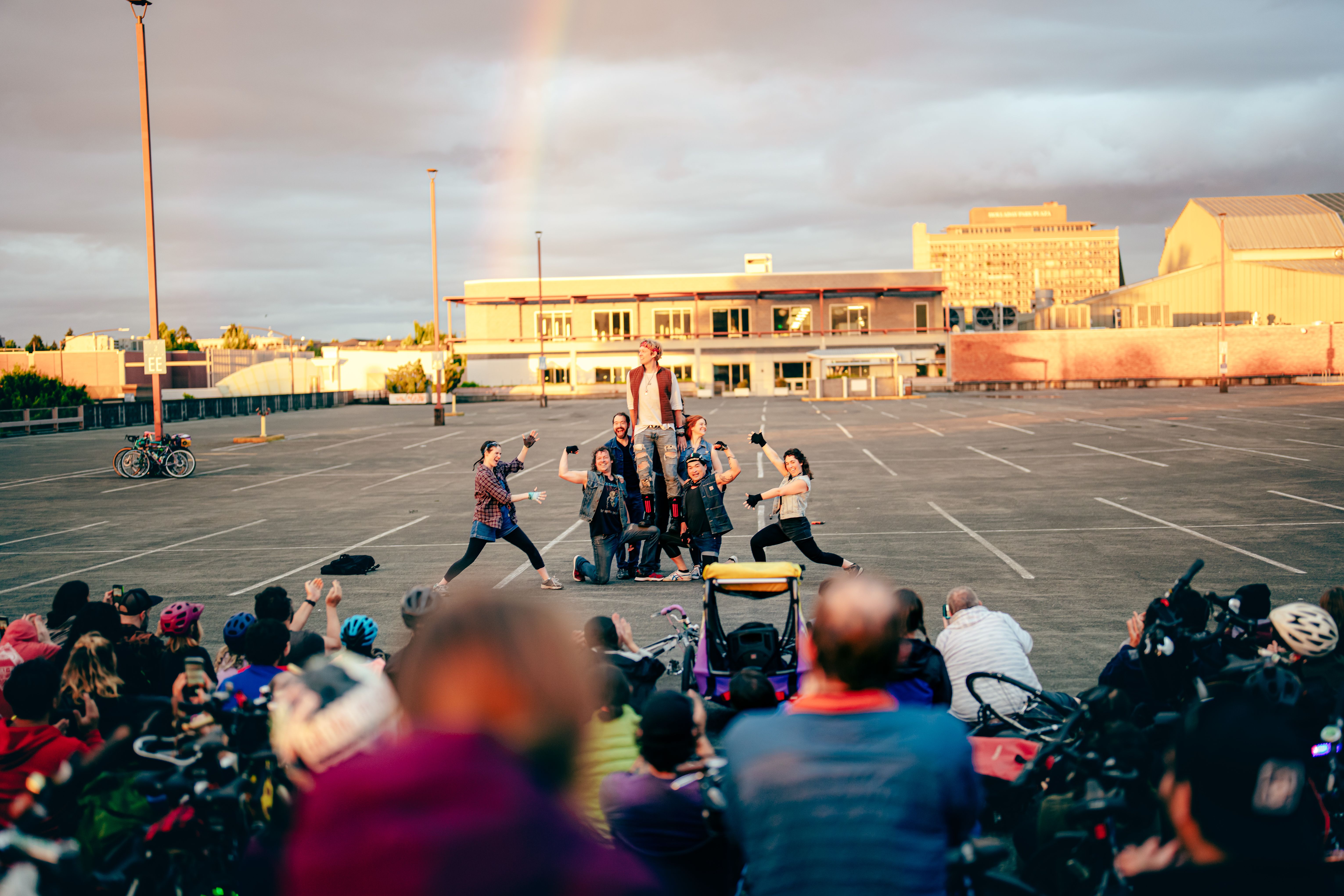 People perform a play in a parking lot with a rainbow in the sky behind.
