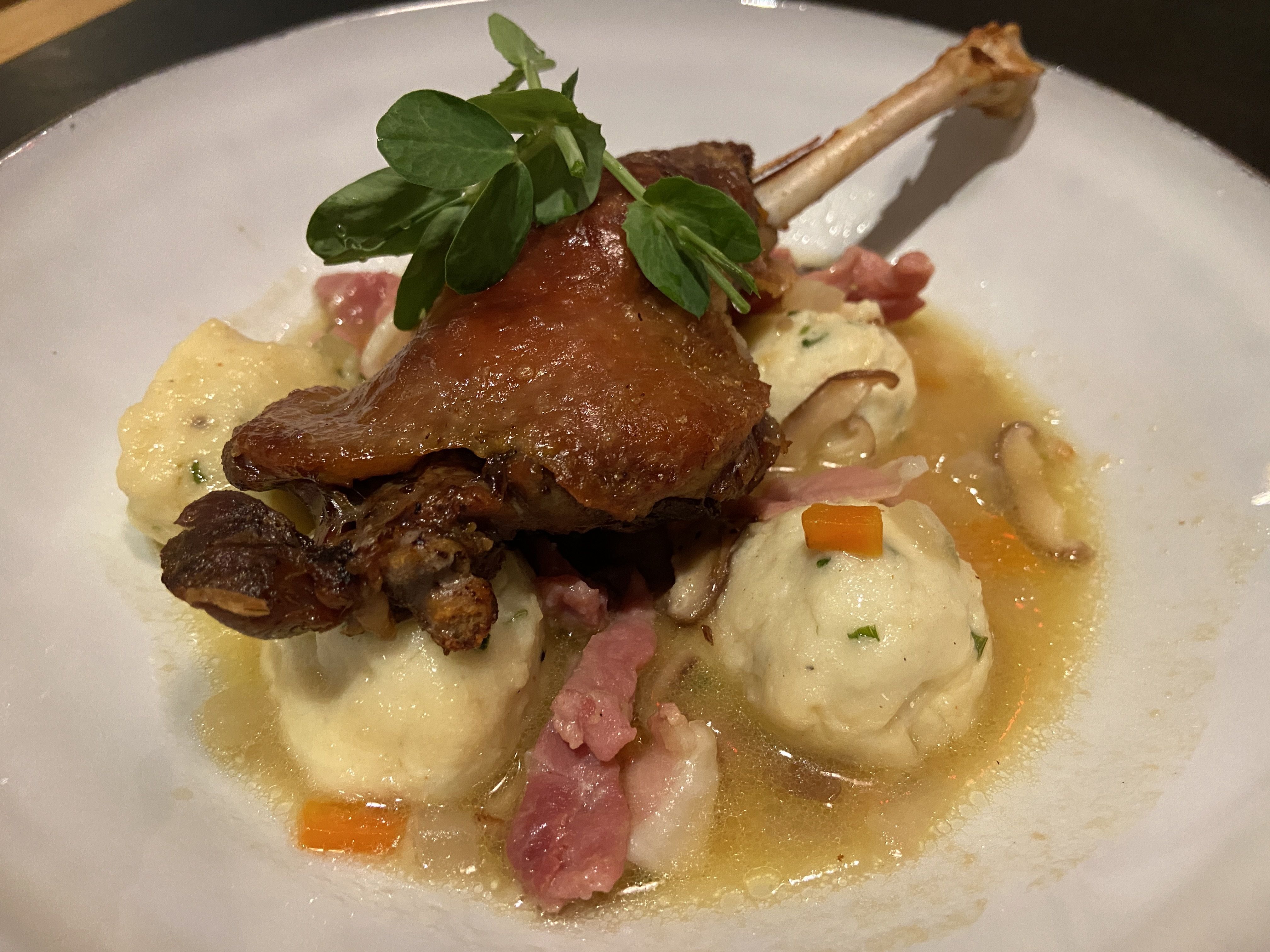 Plate with crispy duck leg on dumplings, garnished with pea shoots, served with bacon, mushrooms, and carrots in a light broth.