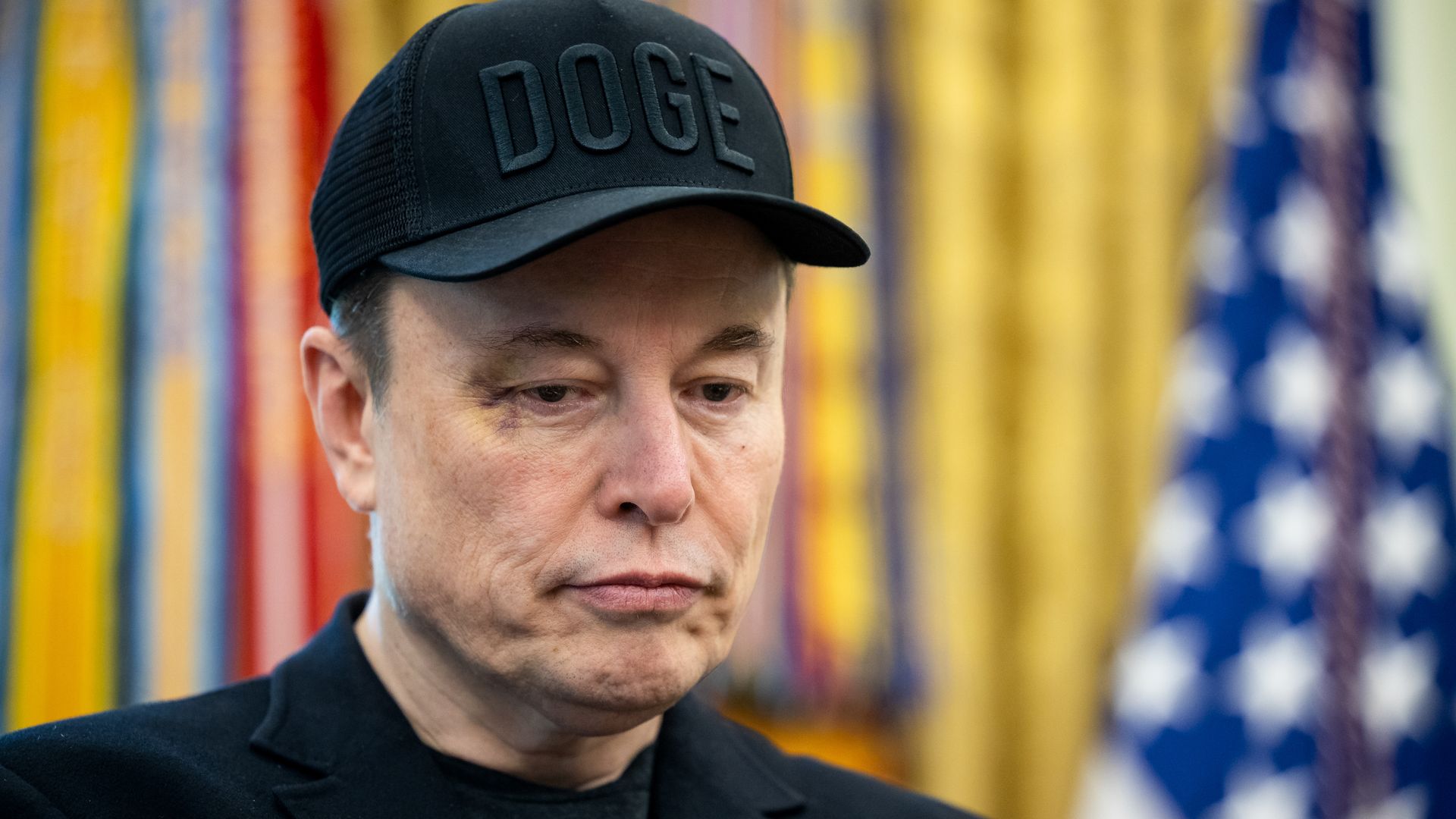 Man wearing a black cap with "DOGE" in uppercase letters, black jacket, with a serious expression; background includes blurred colorful vertical ribbons and a partially visible U.S. flag.