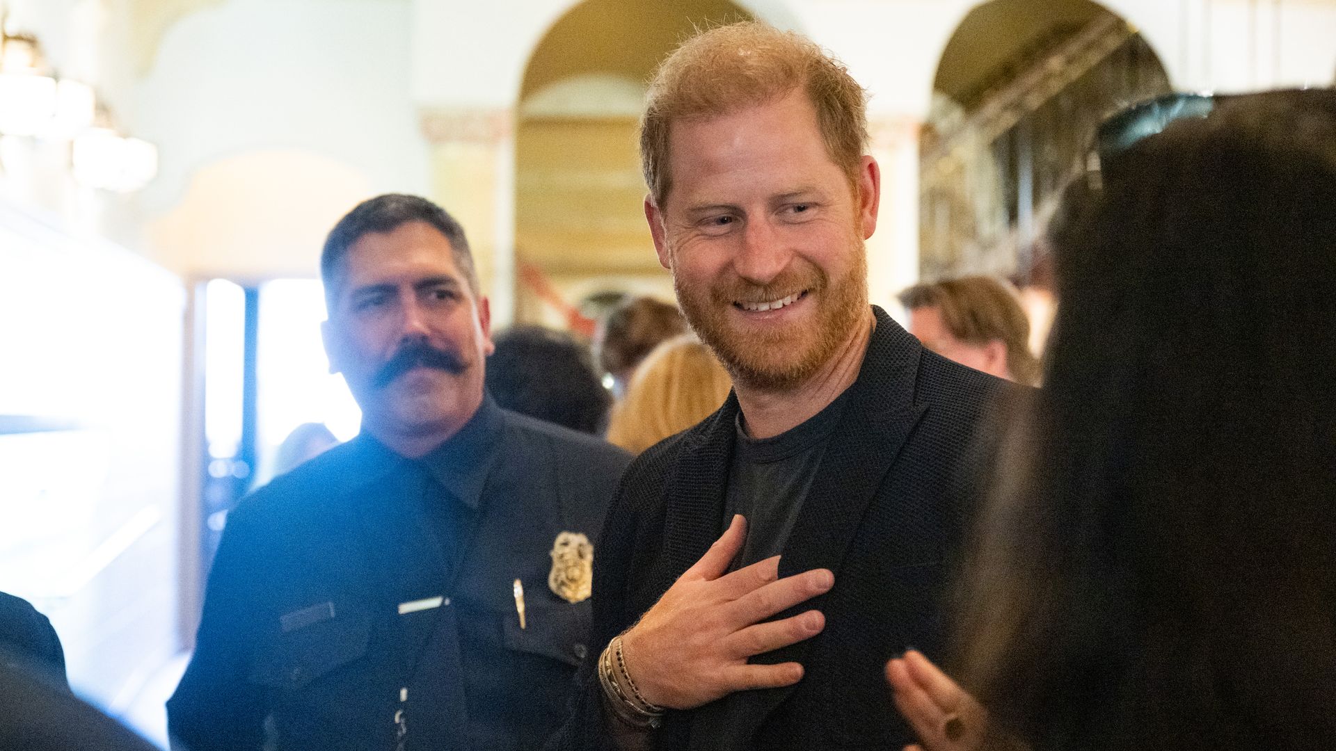 Prince Harry smiles while talking to a person in a room with white walls and vaulted ceilings, with a police officer behind him looks on.