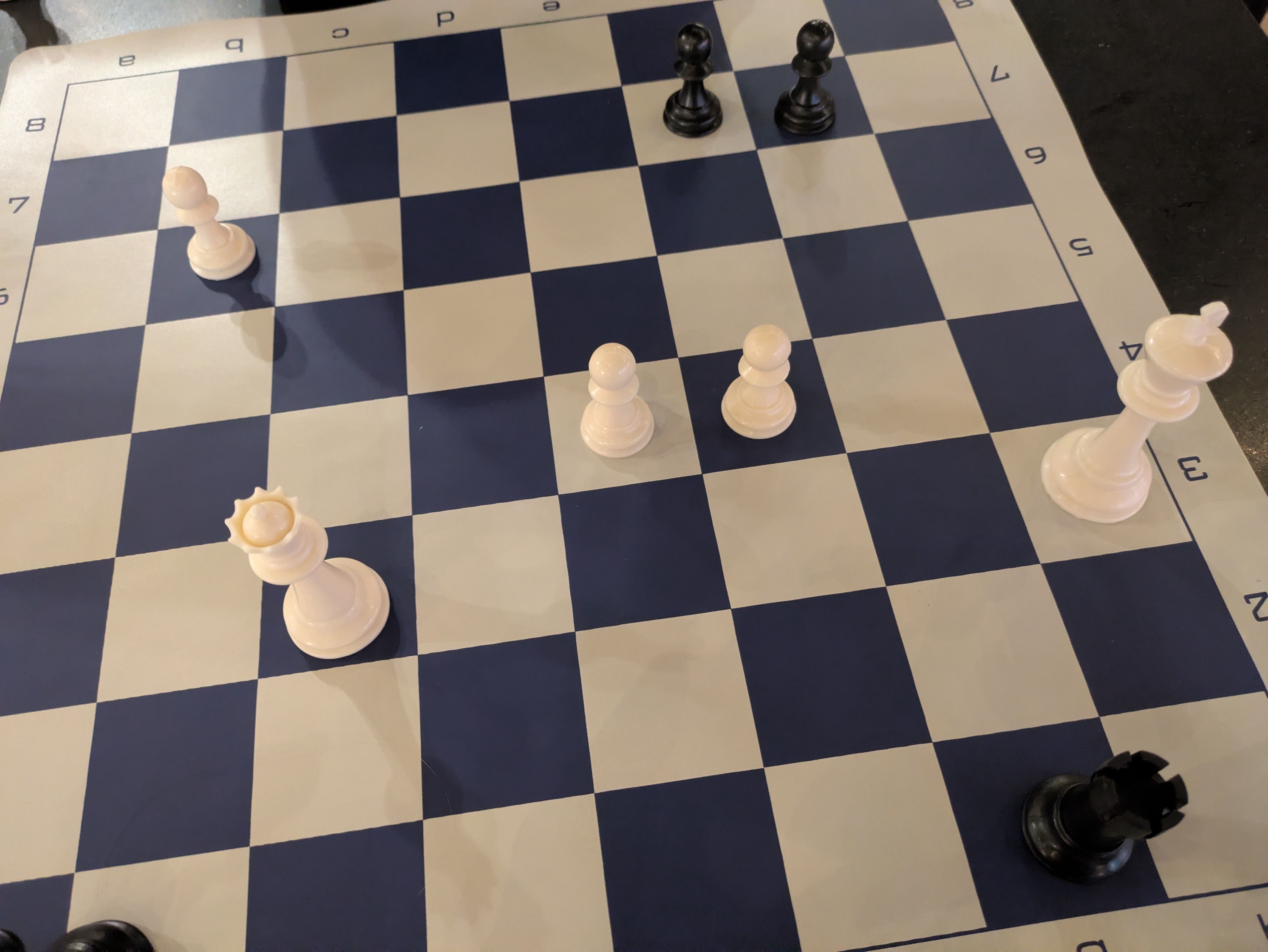 Top-down view of a chessboard with white and black pieces scattered: two black pieces near the top center, several white pawns along the bottom, and a tall white piece in the lower-right corner.