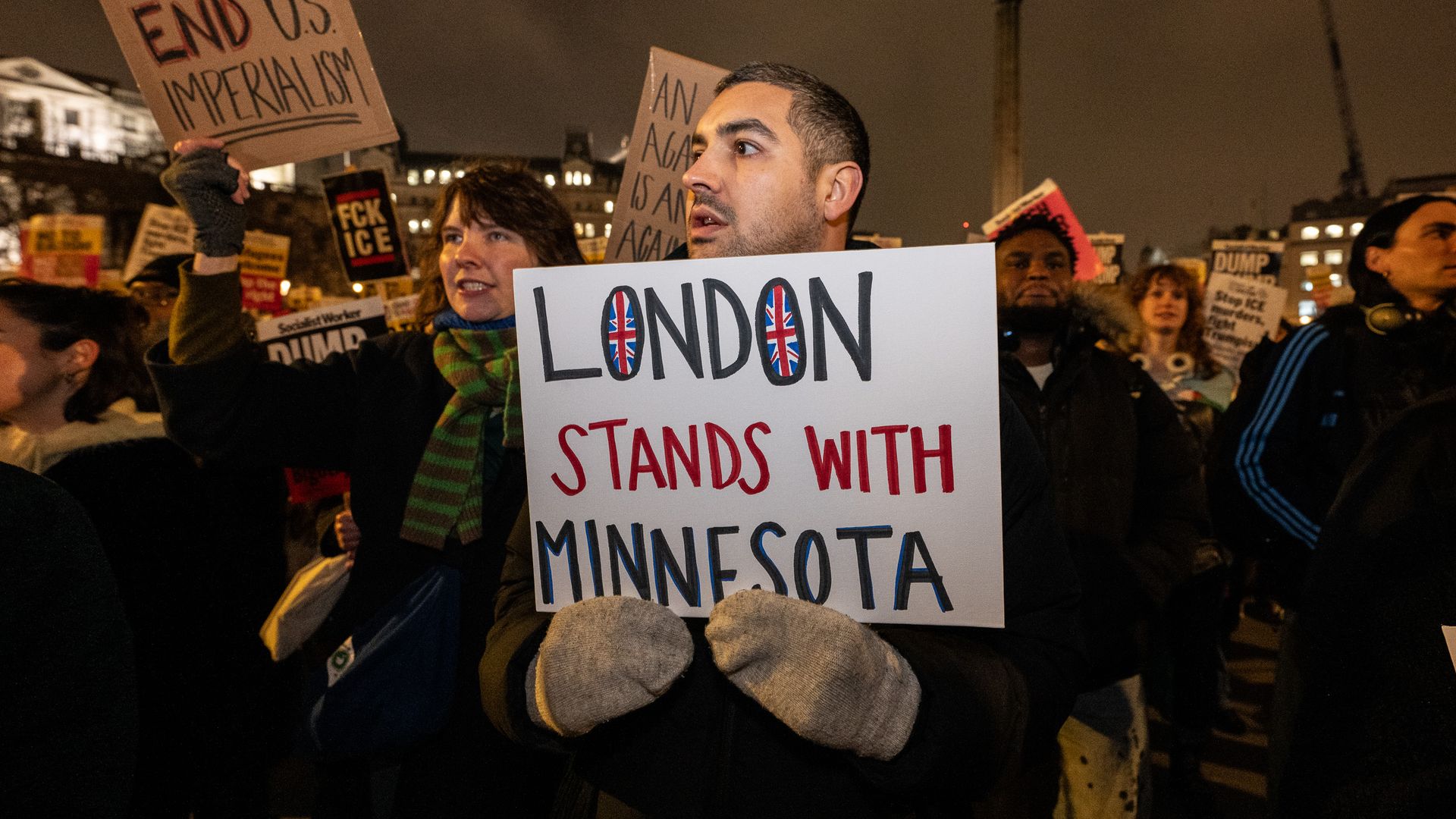 Night protest in London with diverse crowd holding signs including one saying "London stands with Minnesota" with British flags inside letters, and others opposing U.S. imperialism.