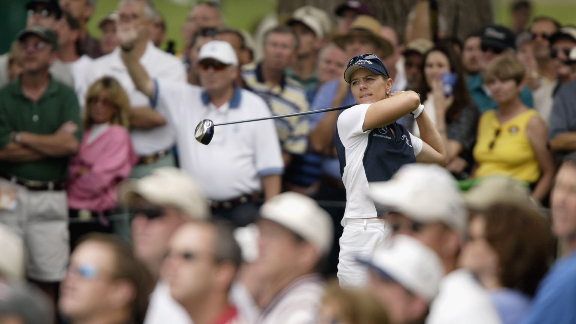 A woman swings her golf club with people watching her from behind