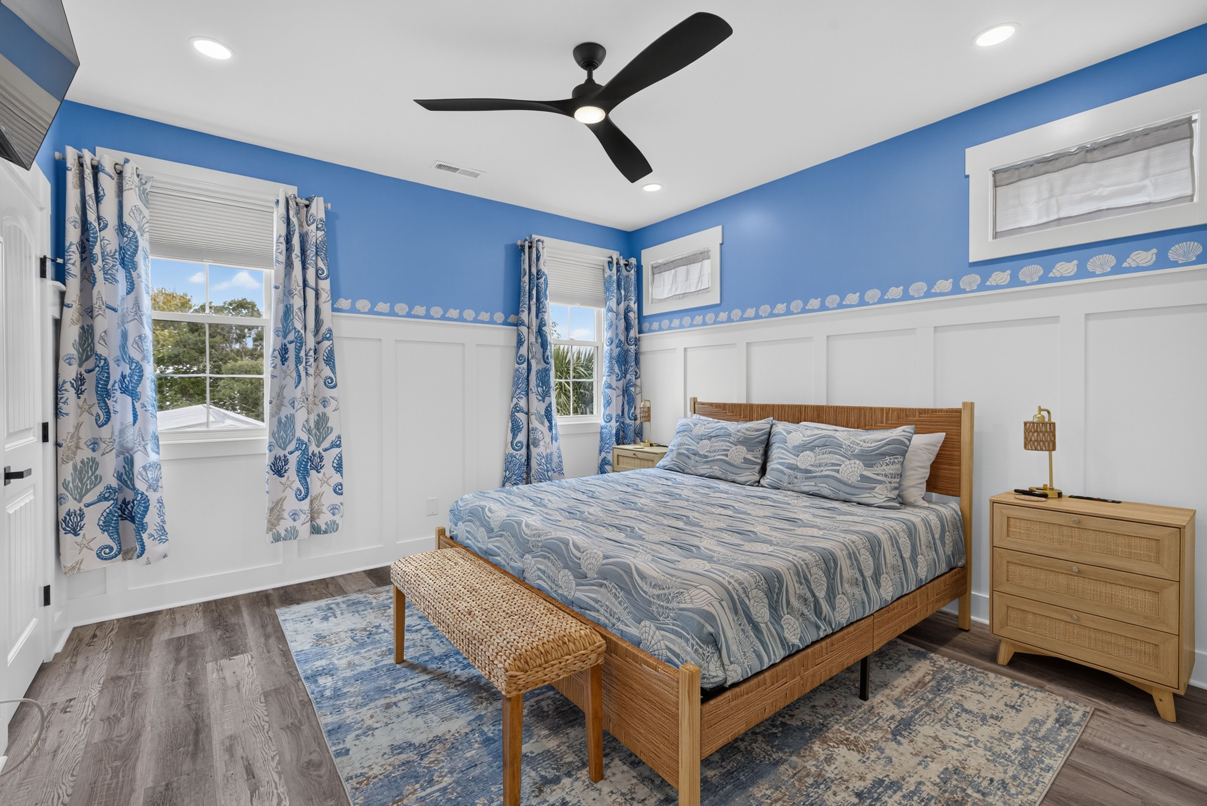 Coastal blue-and-white bedroom with white wainscoting, seashell border, wicker bed, blue patterned bedding, matching curtains, two nightstands, ceiling fan, and windows with blinds.