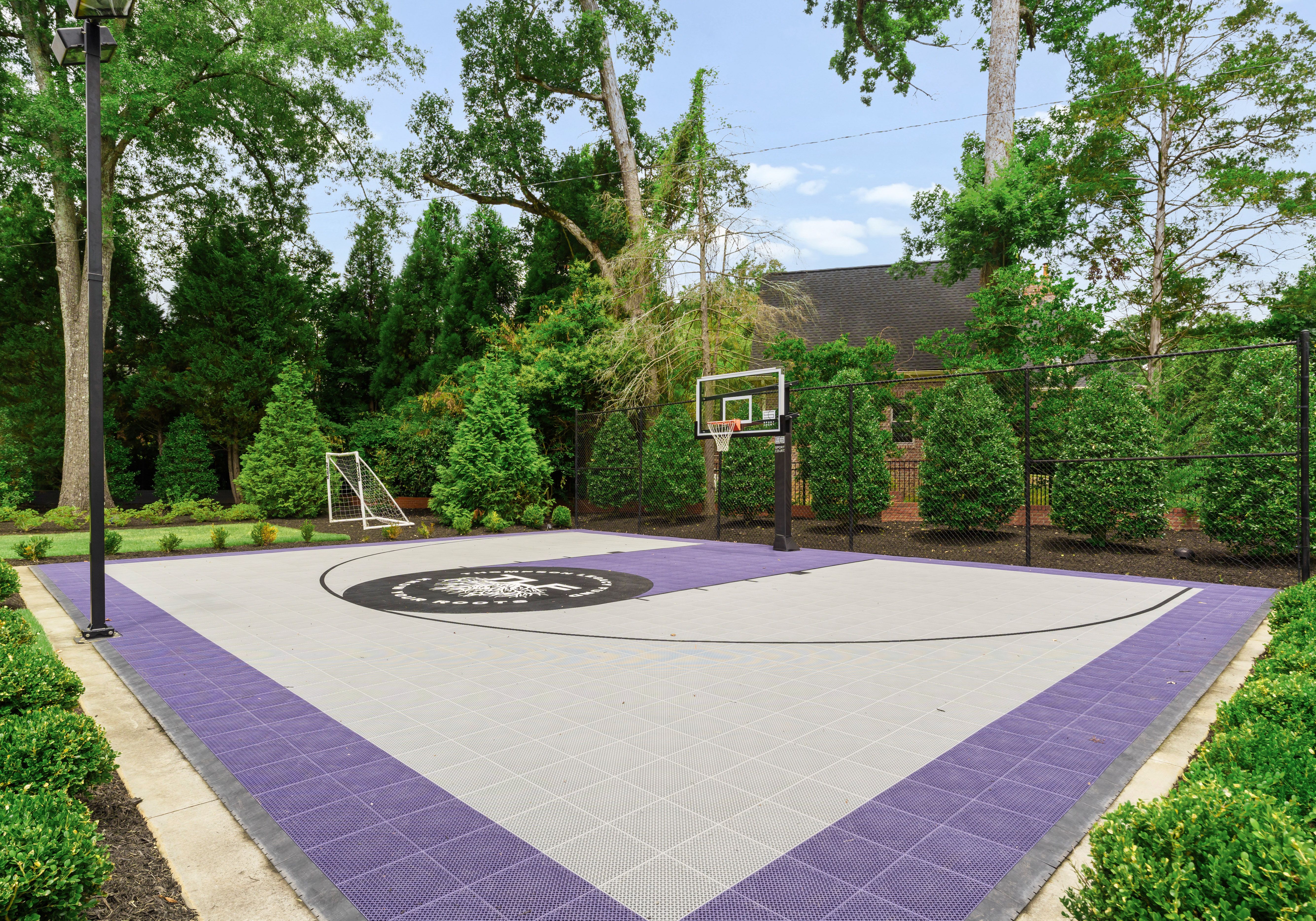 Outdoor basketball court with purple and gray tiles, black basketball hoop, and small soccer goal, surrounded by green trees and bushes under a blue sky.