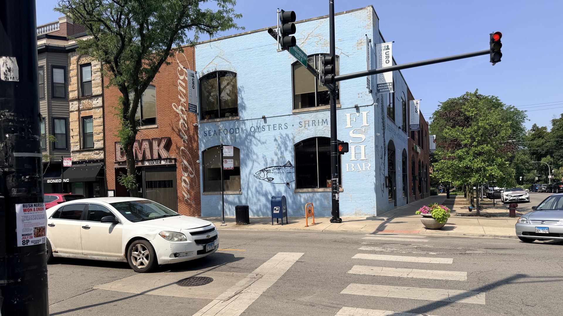 Sunny street scene with a blue building featuring a FISH BAR mural and fish graphic; signs advertise SEAFOOD, OYSTERS, SHRIMP, plus a red brick building, traffic lights, banners, and parked cars.