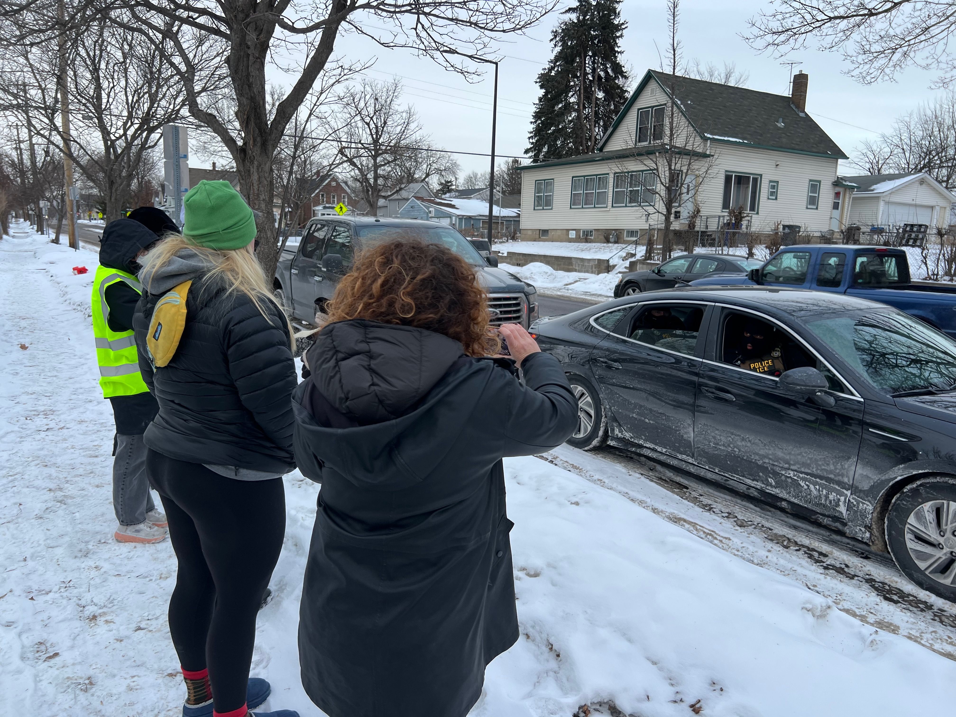 Four people standing on snowy sidewalk near bare trees, one wearing neon yellow vest, facing a black car with masked officers labeled ICE inside, in a residential snowy neighborhood.