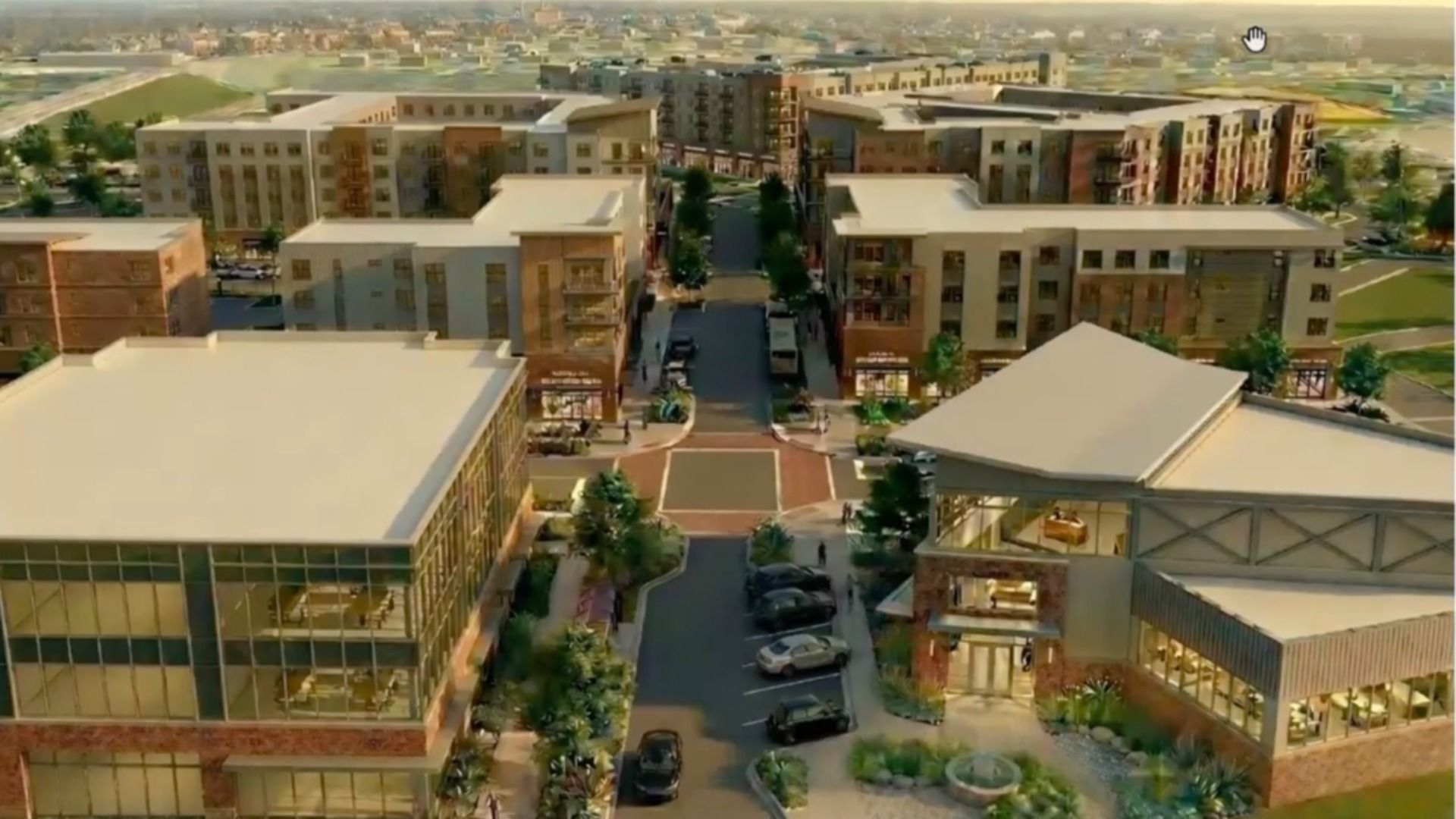 Aerial view of a modern mixed-use development with beige and brick multi-story buildings along a central street of shops, trees, and parked cars, with a desert landscape in the distance.