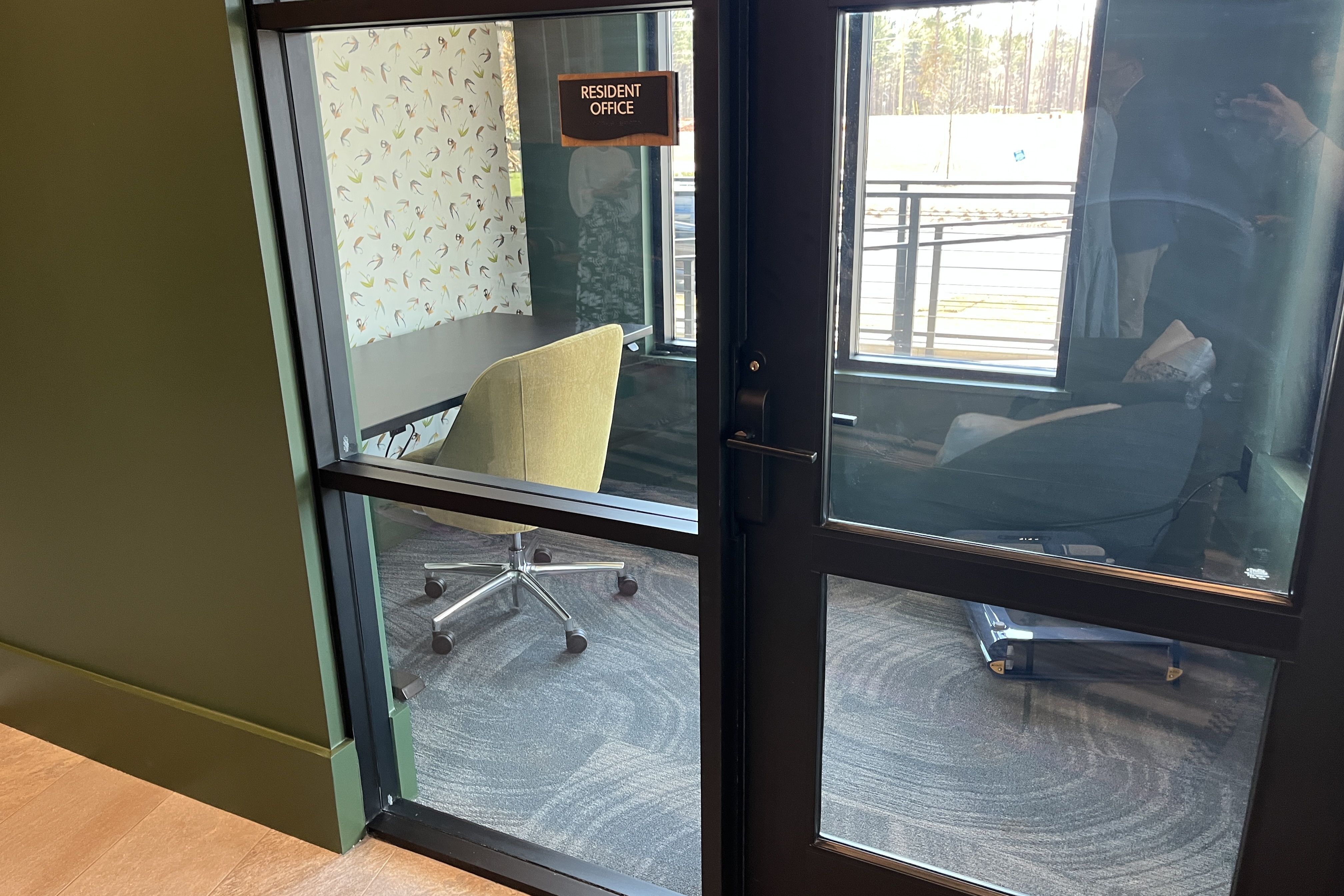 Inside a glass-walled office labeled "Resident Office": a yellow swivel chair at a gray desk, leaf-pattern wallpaper, green walls, and a view of railing and trees outside.