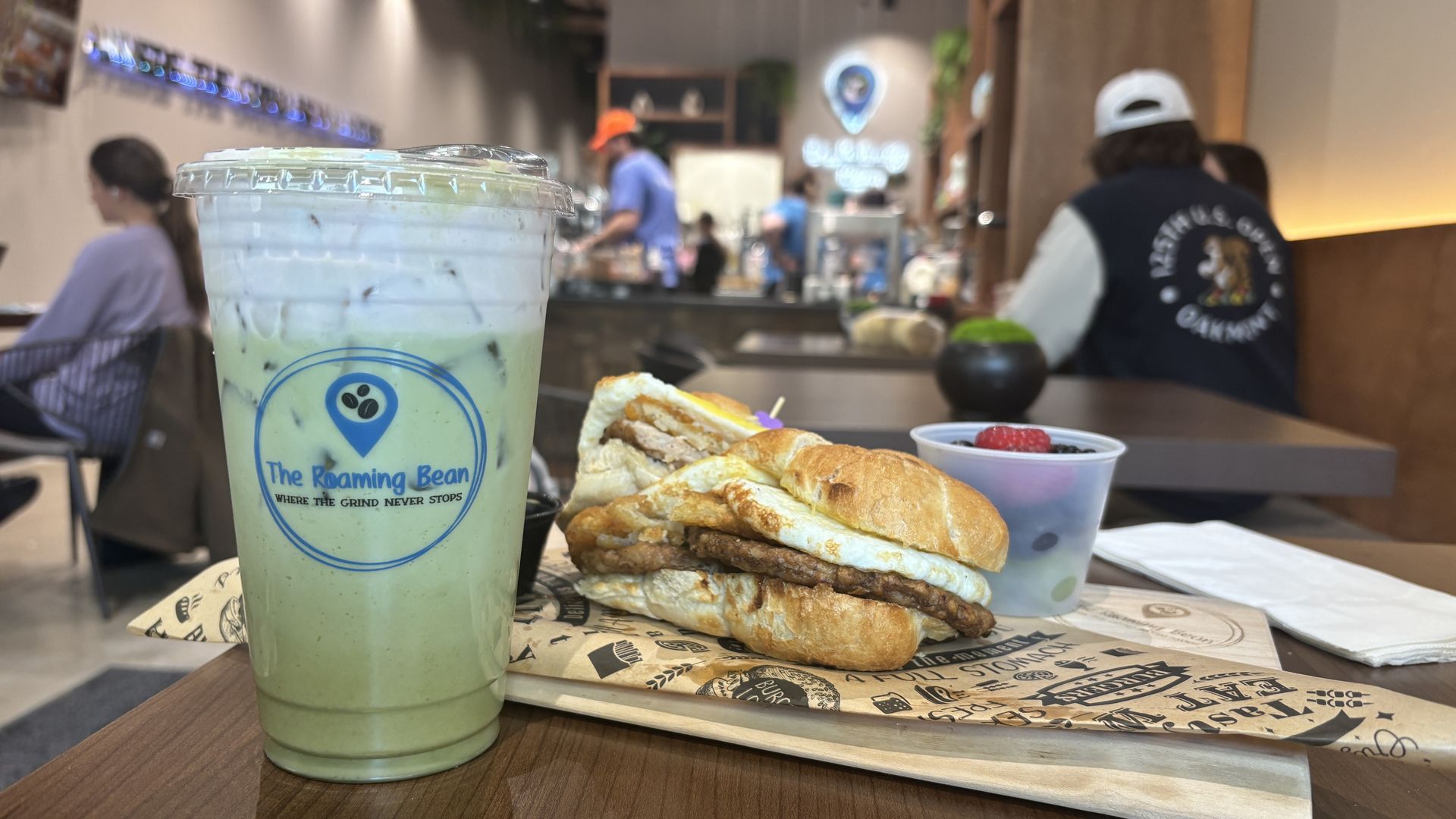 Iced matcha green tea drink in a clear plastic cup with "The Roaming Bean" logo, a breakfast sandwich with egg and sausage on a wooden tray, and a small cup of mixed berries on a table in a café.