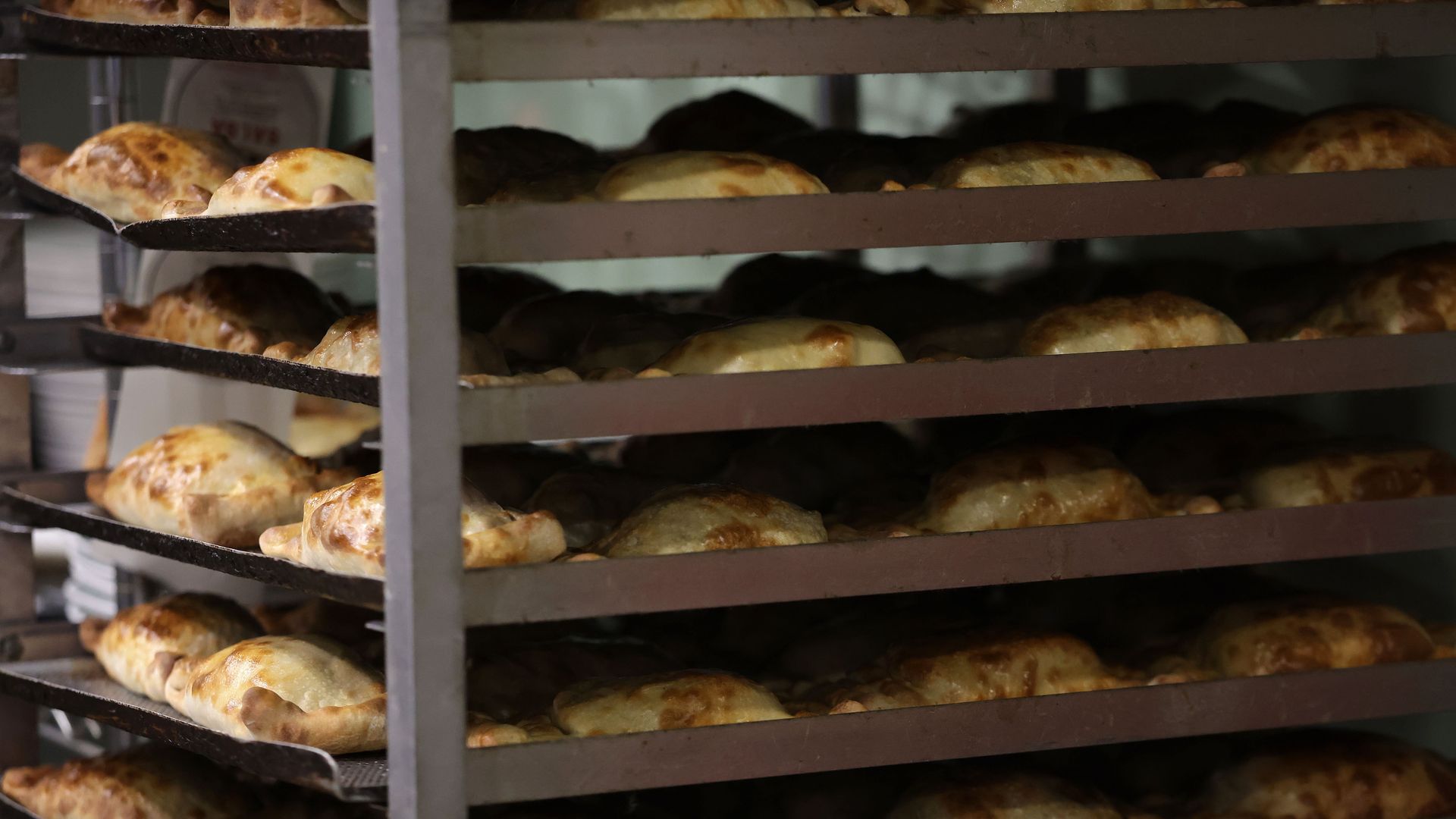 Rows of golden-brown empanadas on metal baking racks in a bakery, stacked on multiple trays, warm and freshly baked.