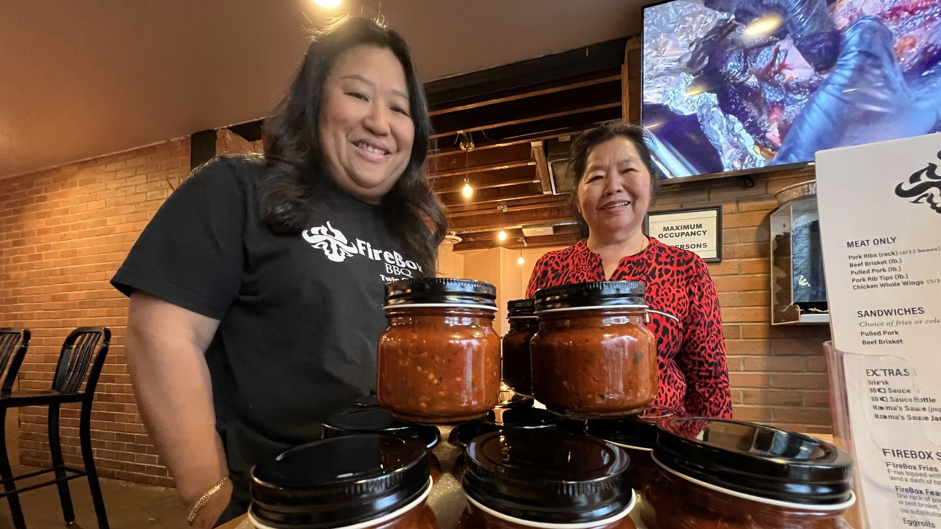 Two Asian women smile for the camera with jars of red pepper sauce in the foreground. The woman on the left wears a black T-shirt that reads 'Firebox BBQ,' the woman on the left is in red.